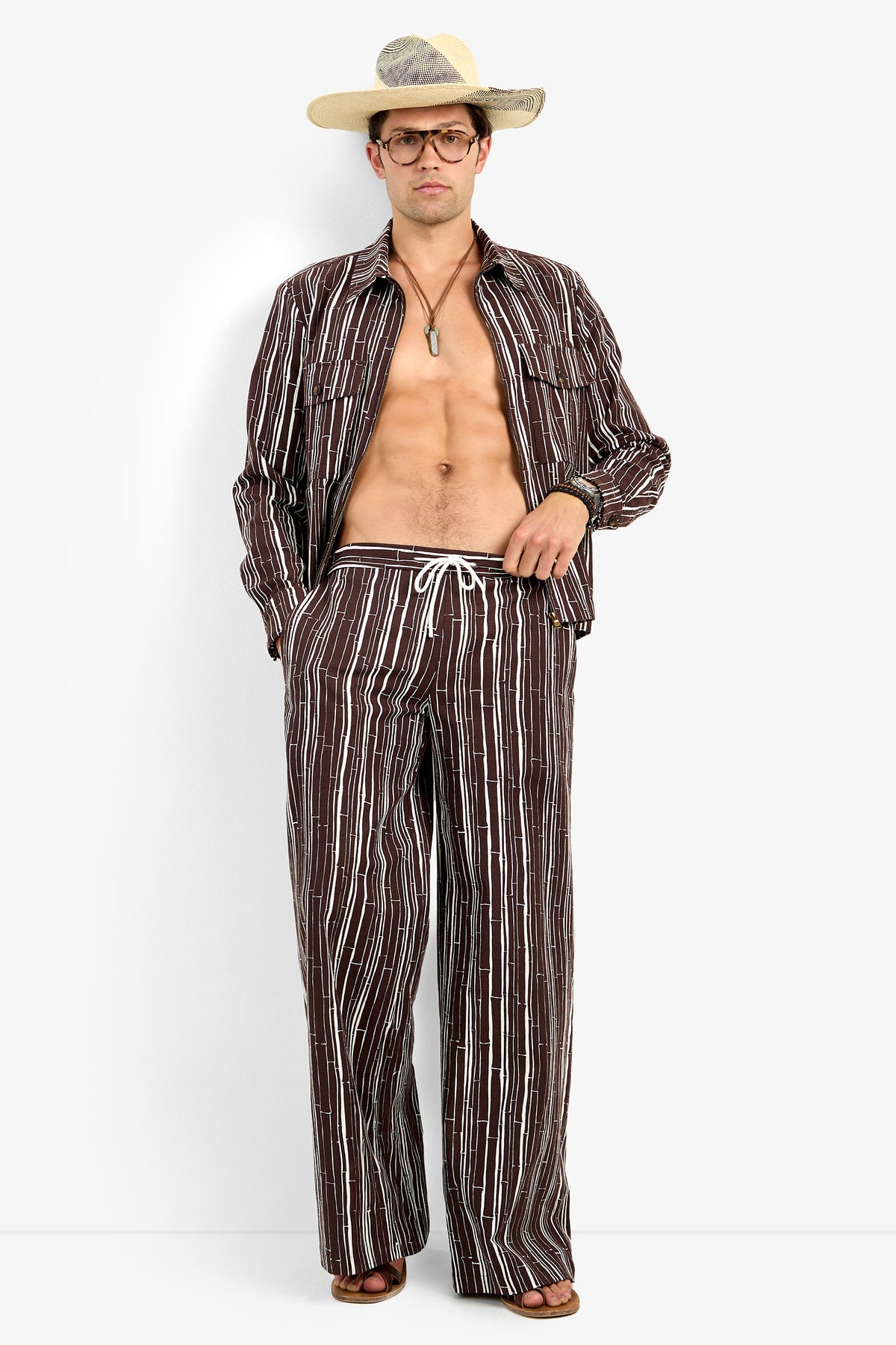 A man stands against a plain white background wearing the Arturo Jacket, an open brown striped shirt, paired with matching drawstring pants, a wide-brimmed hat, glasses, sandals, and a necklace.