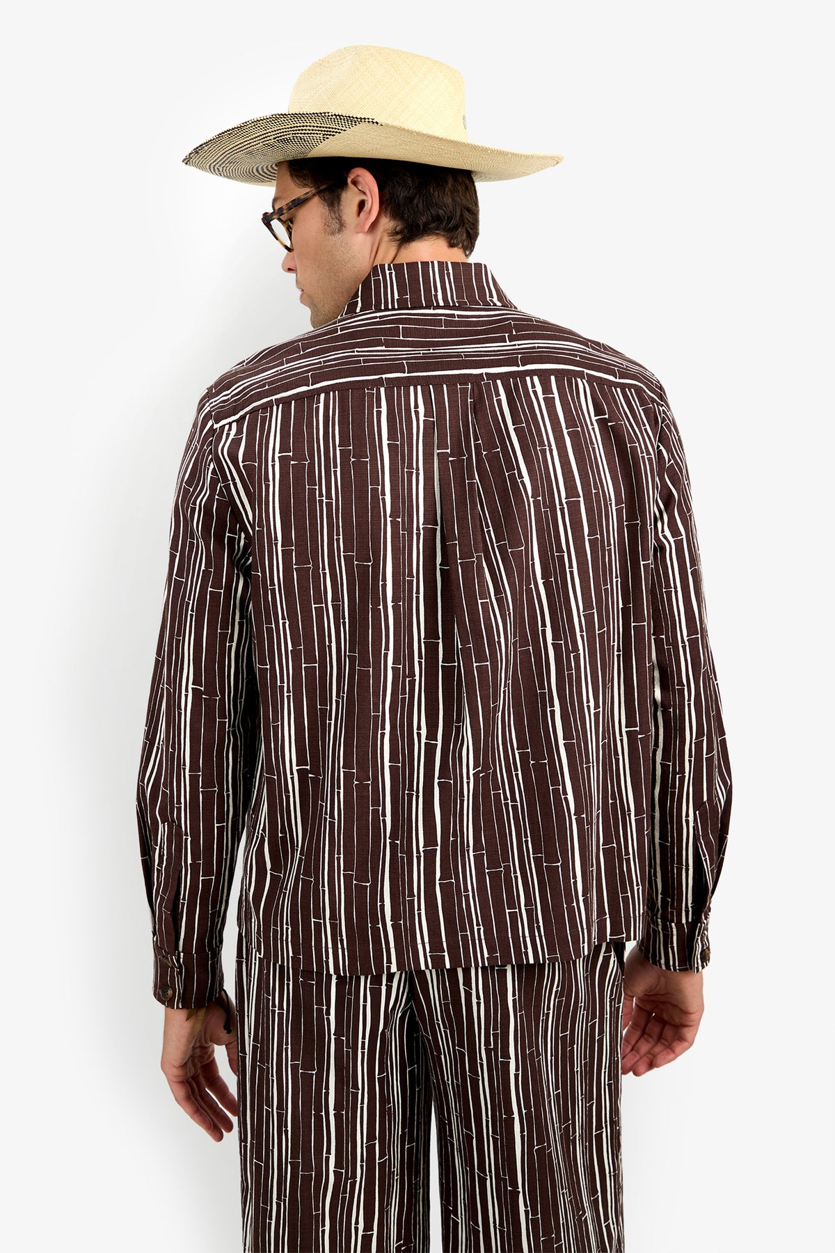 A person in the Arturo Jacket and matching brown pants with white vertical stripes, straw hat, and glasses stands facing away, showing the back of the outfit against a plain background.