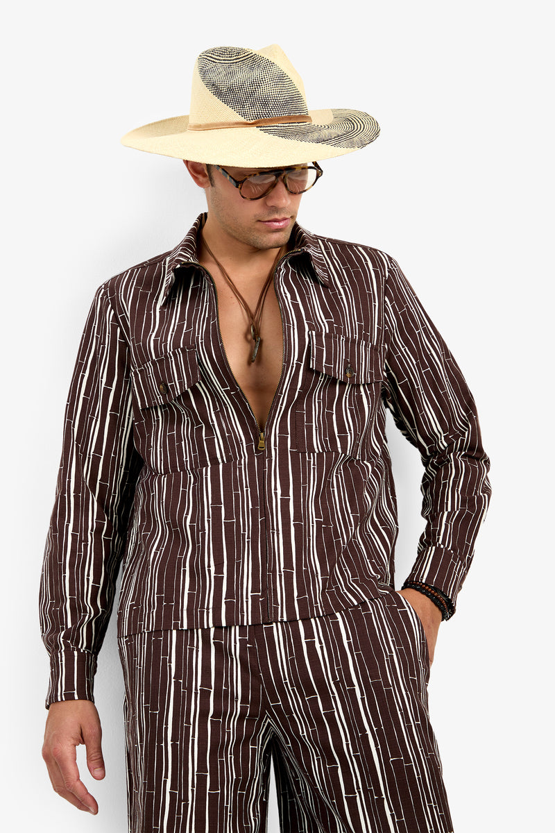 A man wears the Arturo Jacket with matching pants, accessorized with a beige wide-brim hat, black-rimmed glasses, a necklace, and a bracelet, posing stylishly with one hand in his pocket.