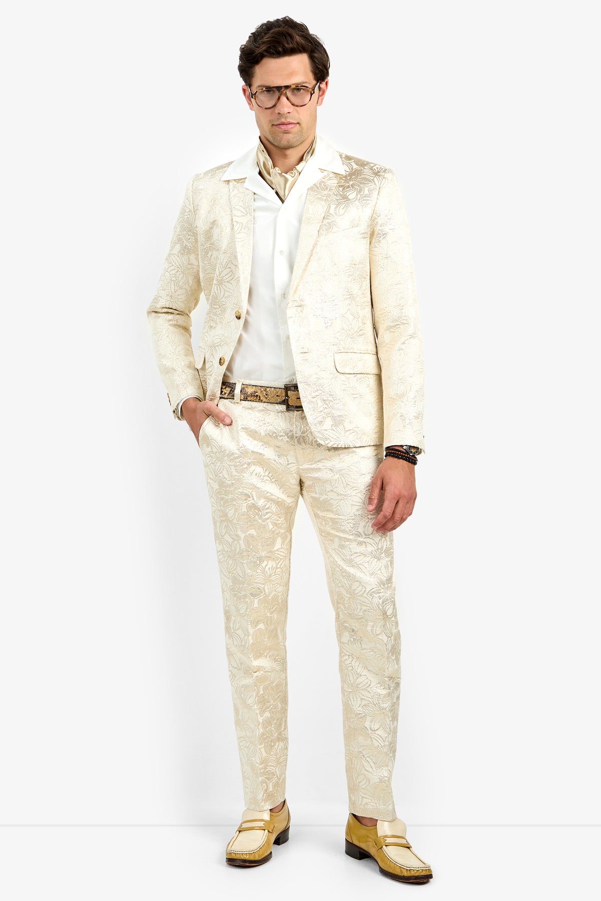 A man with short brown hair, glasses, and a neutral expression models the Thurston Blazer—a cream-colored, patterned suit worn over a white shirt with a gold belt and yellow shoes—against a plain white background.