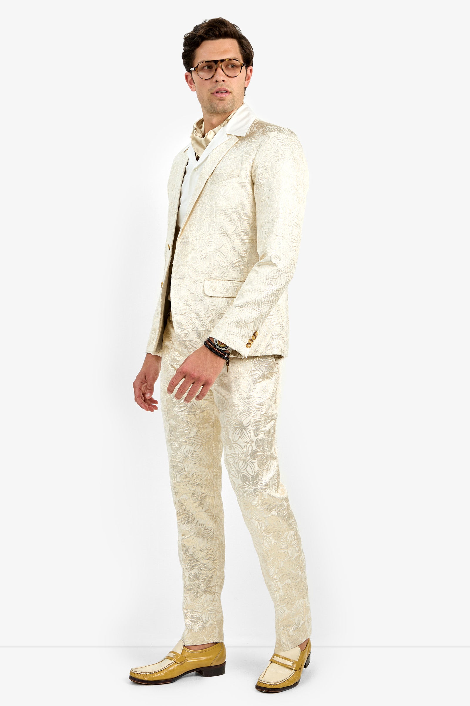 A man wearing glasses and the Thurston Blazer—a cream-patterned suit—with a white shirt, tan shoes, and bracelets poses against a plain white background, standing slightly to the side.