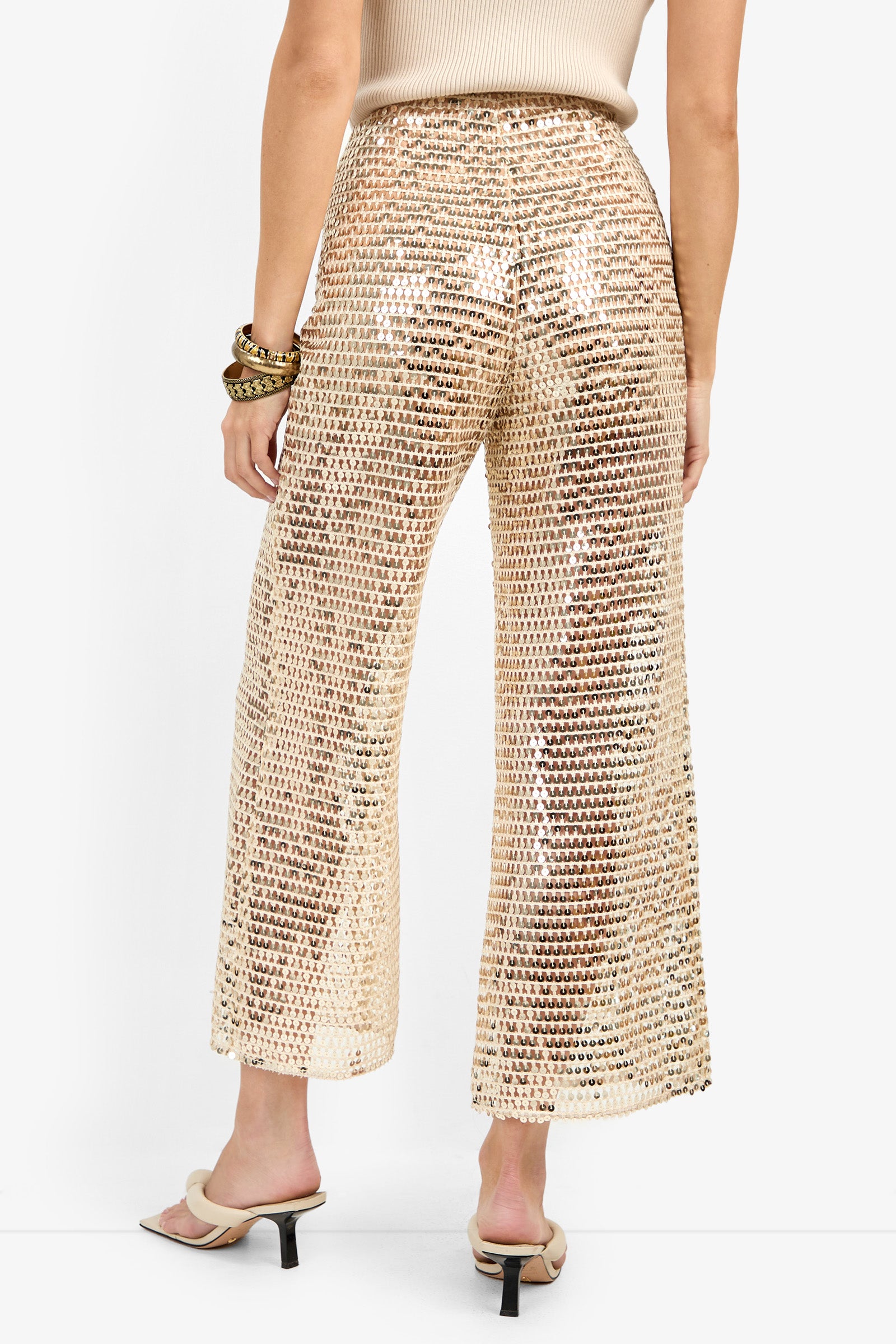 Wearing the Dariya Pant—high-waisted, wide-leg, gold sequin mesh pants—with a beige sleeveless top, heeled sandals, and chunky bracelets, a person stands with their back to the camera against a plain background.
