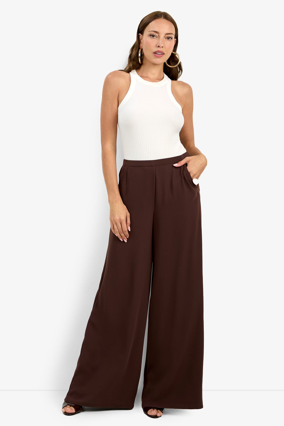 A woman poses against a white background wearing the Aloness Pant—high-waisted, wide-leg brown pants—paired with a sleeveless white ribbed top, one hand in her pocket and the other by her side.