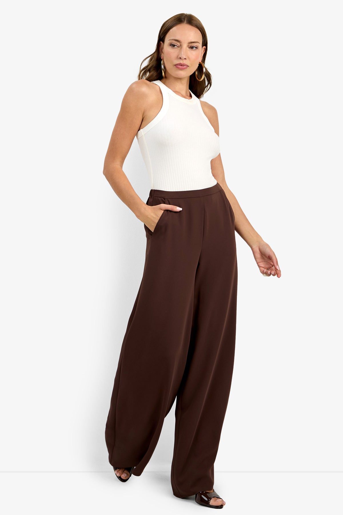 A woman stands against a white background wearing a sleeveless white ribbed top and Aloness Pant—high-waisted, wide-leg brown pants with pockets. She has loose hair, hoop earrings, and strappy black sandals.