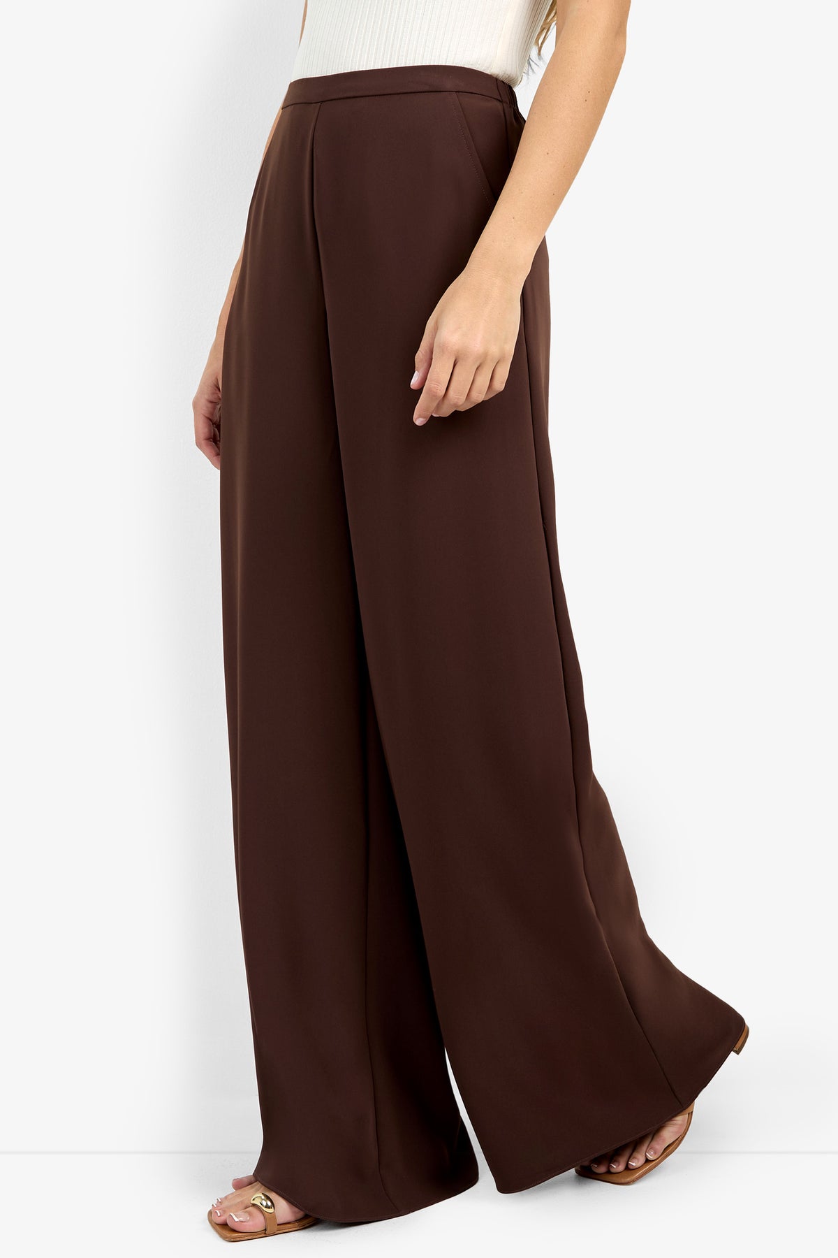 Model wears the Aloness Pant—high-waisted, wide-leg brown pants—paired with a sleeveless white top and flat sandals, shown from the waist down with one leg forward.