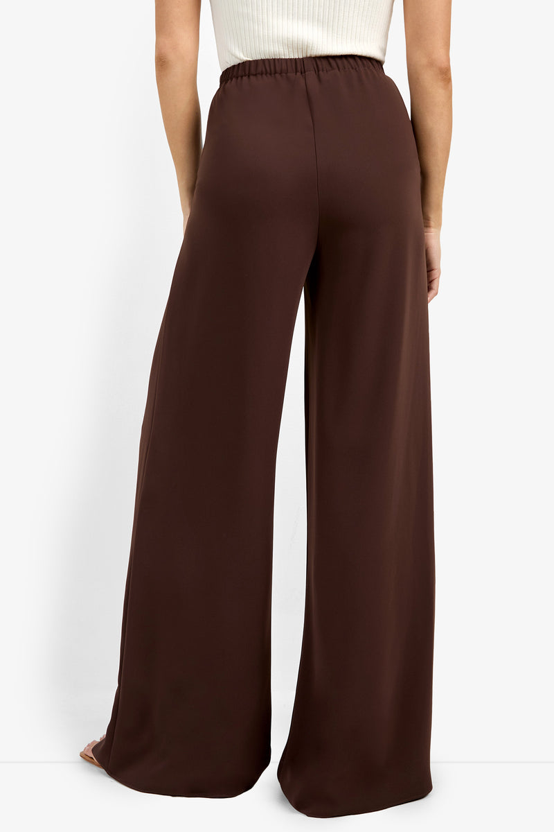 Someone wearing the Aloness Pant—wide-leg, high-waisted, dark brown pants—with a white top, photographed from behind against a white background.