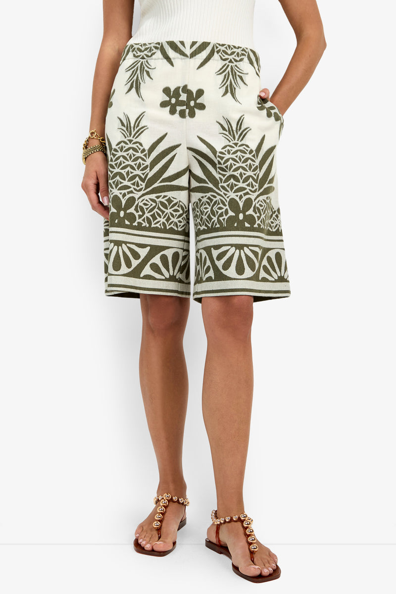 A woman wearing the Estella Short, featuring green and white pineapple and floral prints, paired with a white ribbed top, brown pearl sandals, and a gold bracelet stands with hands in her pockets.