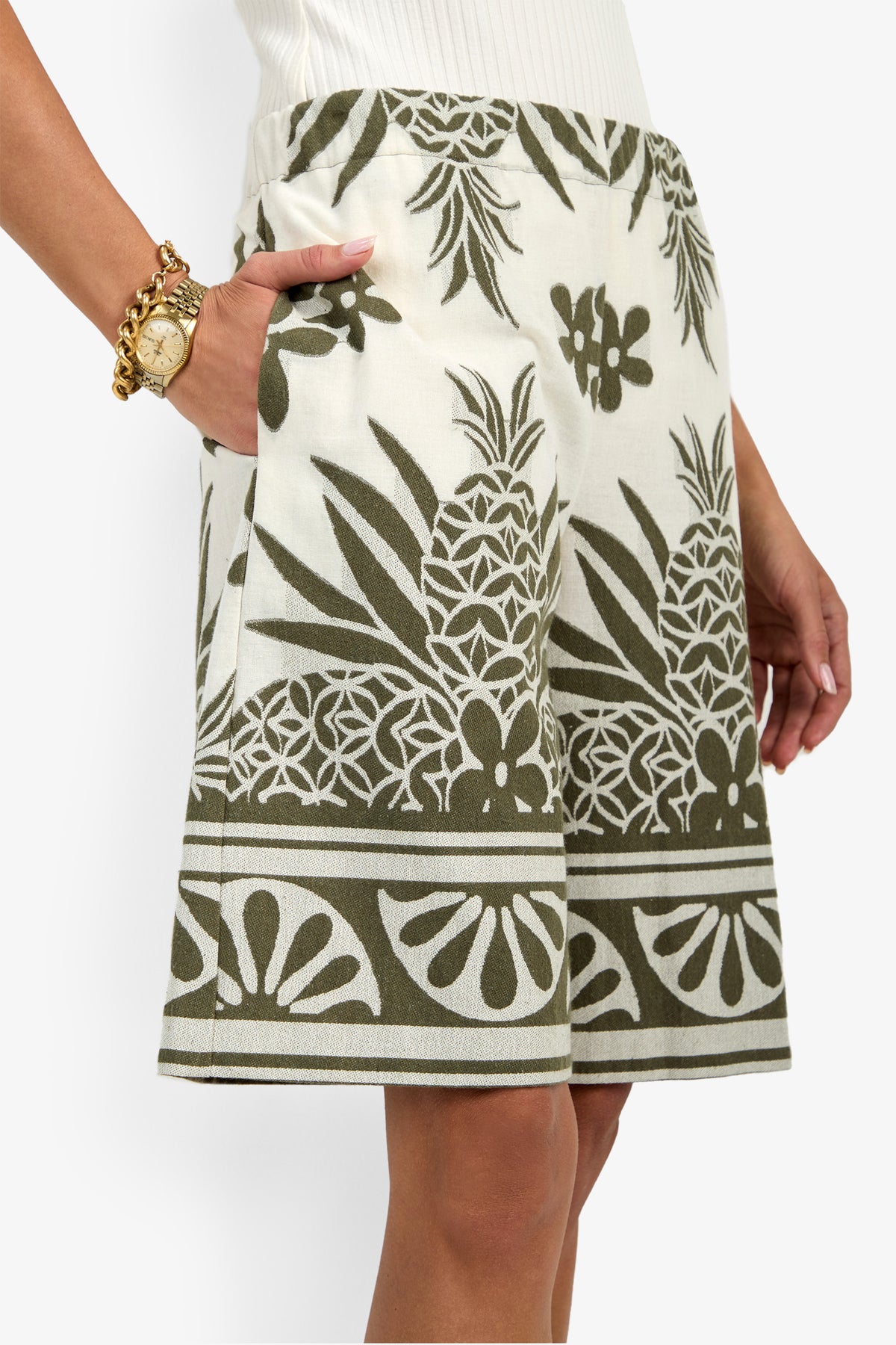 A person wearing the Estella Short—a white short with an olive green tropical pineapple and floral print—stands with one hand in the pocket, paired with a white top, gold bracelet, and watch.