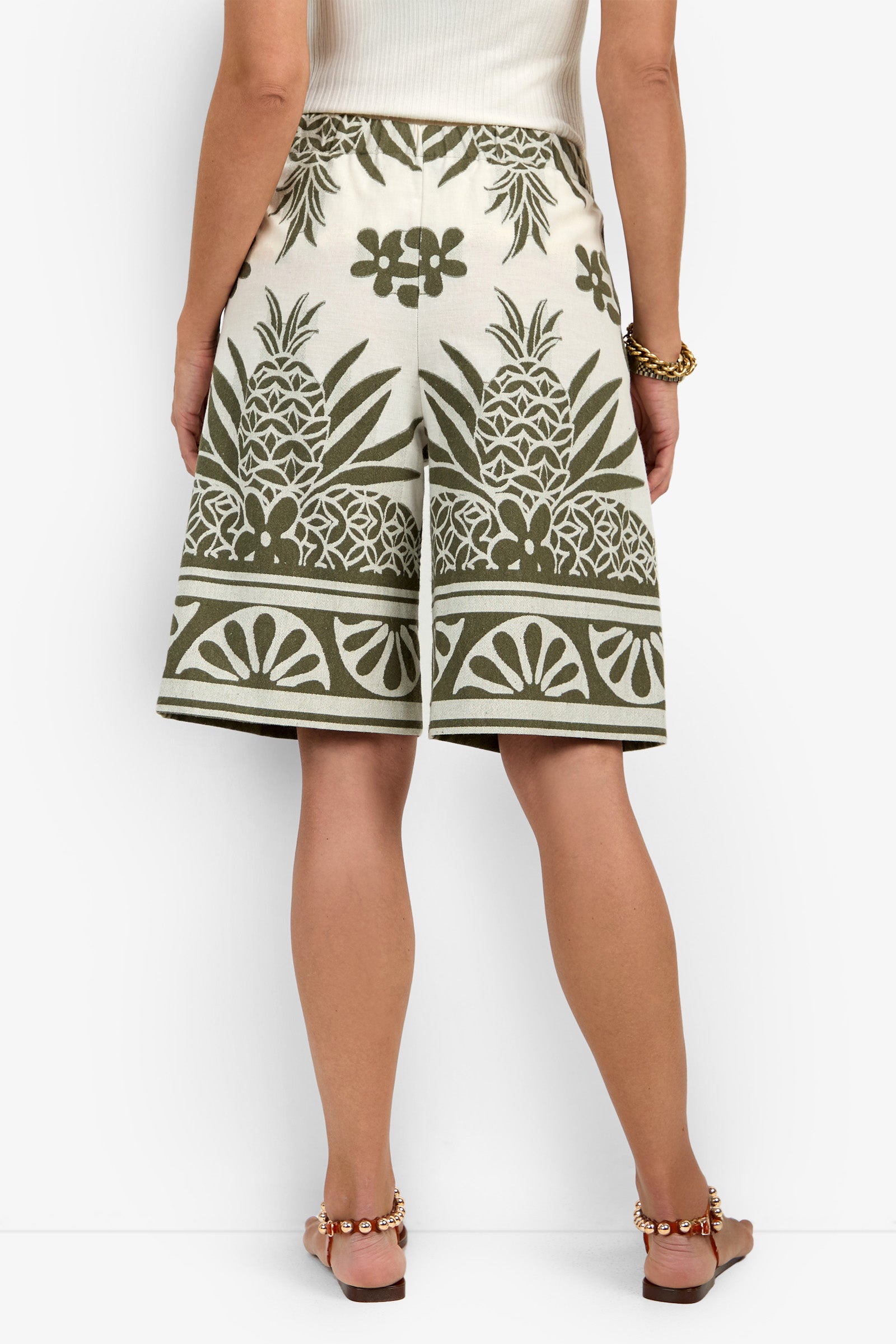 A woman facing away wears the Estella Short in cream with green pineapple and floral prints, paired with a white sleeveless top, brown sandals, and a gold bracelet.