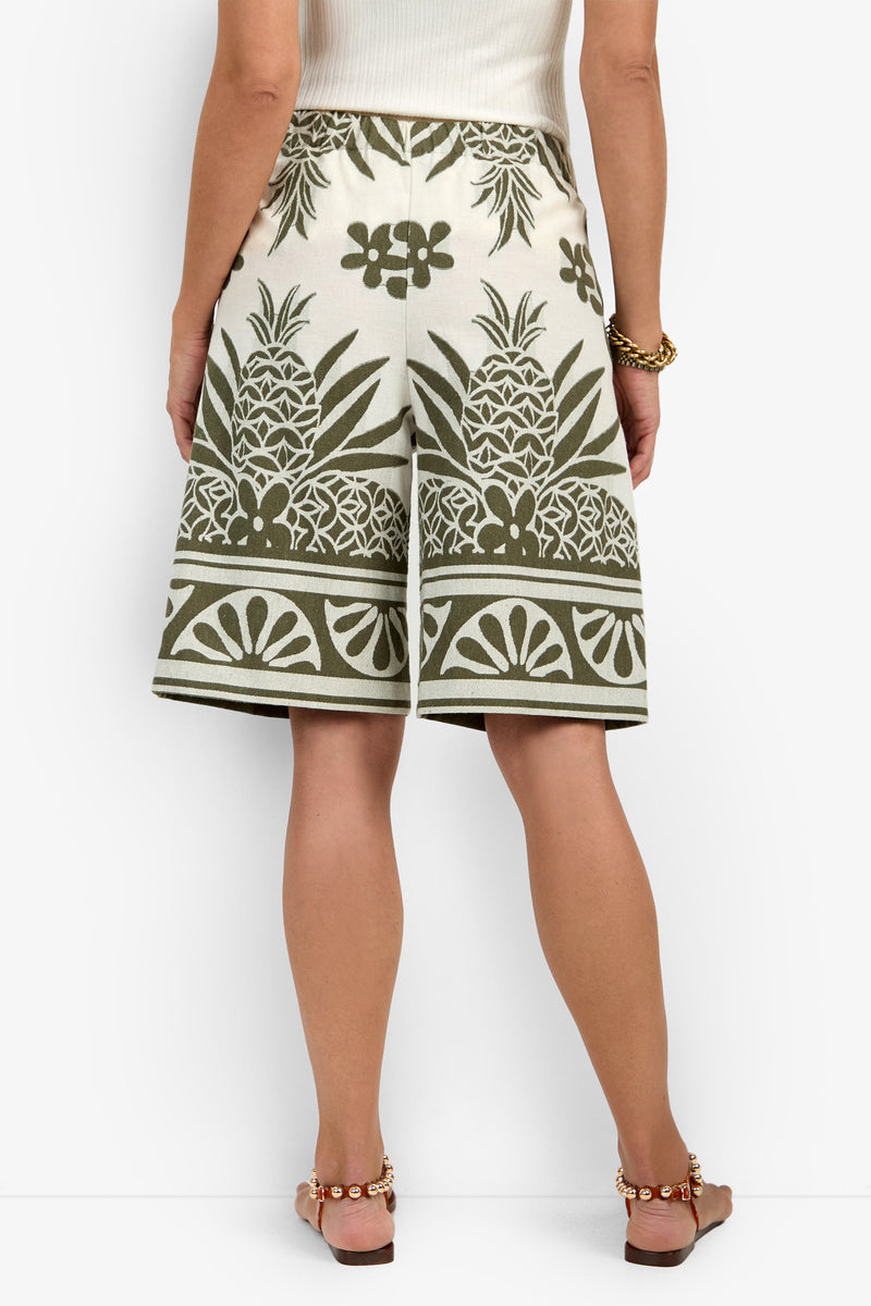 A woman facing away wears the Estella Short in cream with green pineapple and floral prints, paired with a white sleeveless top, brown sandals, and a gold bracelet.