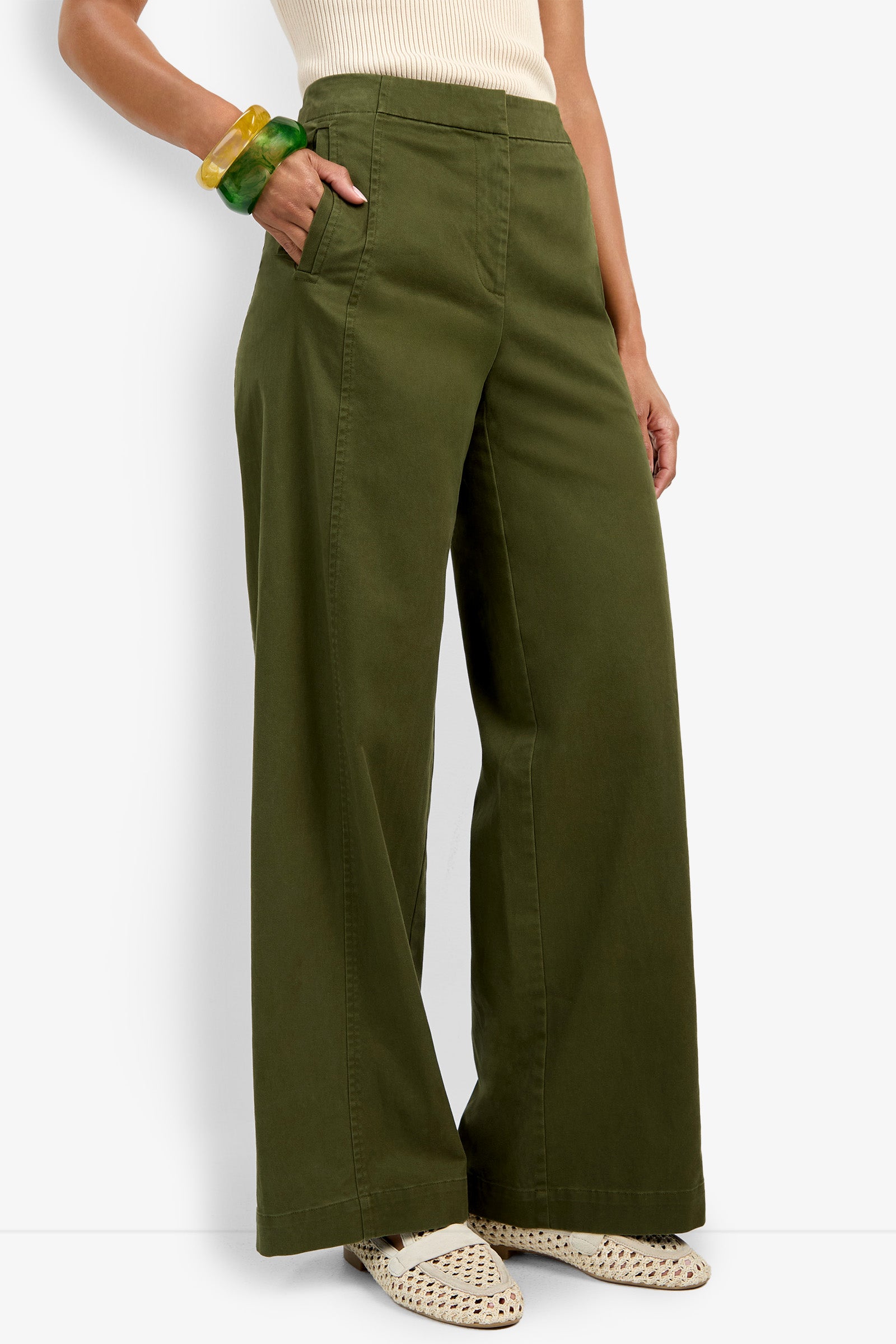 A person wearing the Carum Pant in olive green, paired with a cream sleeveless top, cream woven shoes, and green and yellow chunky bracelets stands against a white background.