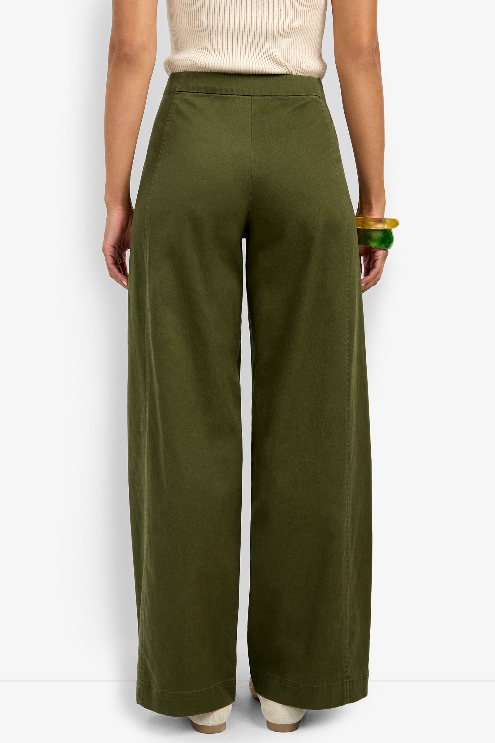 A person wearing the Carum Pant—high-waisted, wide-leg olive green pants—and a sleeveless beige top stands with their back to the camera, accessorized with two chunky bracelets on their right wrist.