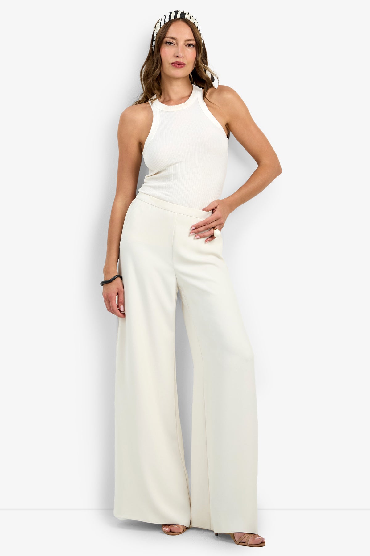 A woman poses with one hand on her hip against a plain background, wearing the Aloness Pant styled with a white sleeveless halter top, open-toe heels, and a black-and-white headband.