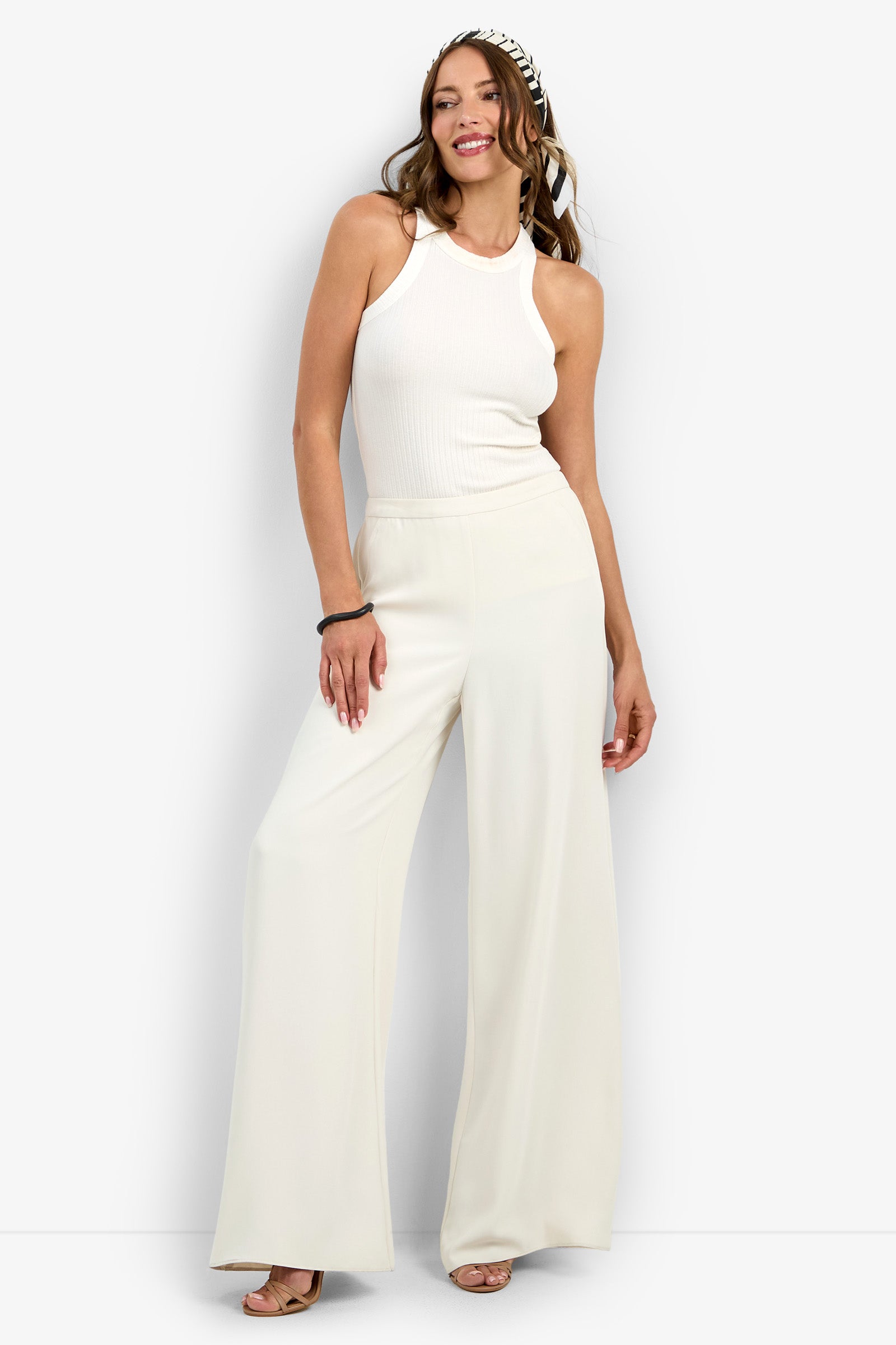 A woman smiles in the Aloness Pant—wide-leg, white pants—paired with a sleeveless white ribbed top, nude heels, long wavy brown hair, a black-and-white headband, and a black bracelet on her right wrist.