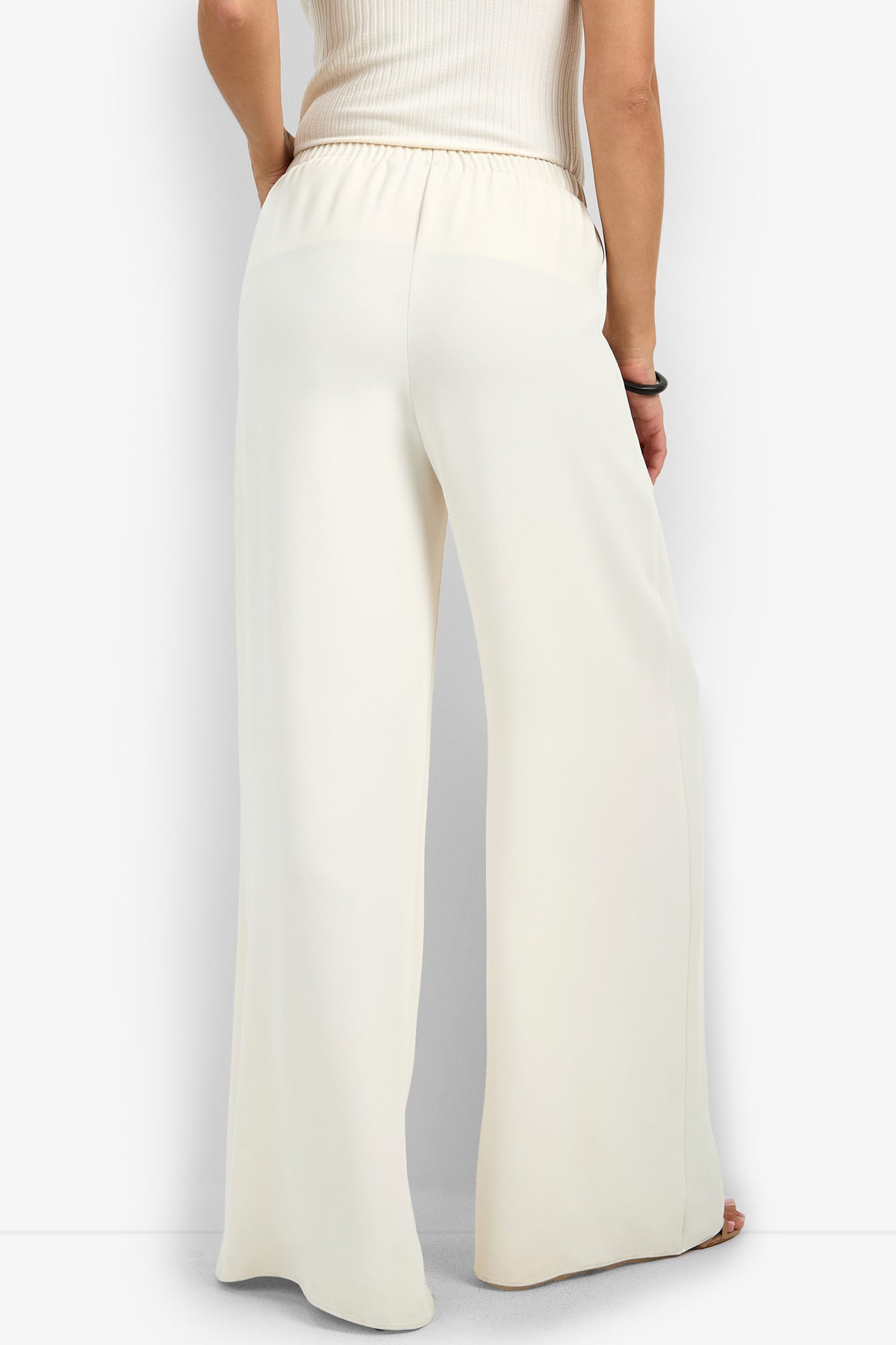 Someone is standing with their back to the camera against a plain background, wearing the Aloness Pant—loose, high-waisted, white wide-leg pants—and a sleeveless ribbed top.