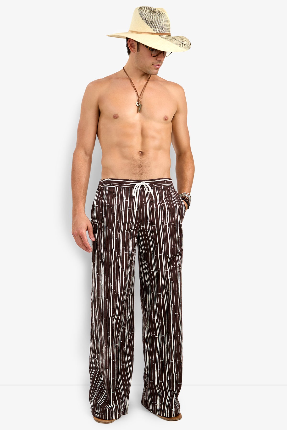 A shirtless man in a straw hat, sunglasses, necklace, wristwatch, and Wakiki Trouser—brown drawstring pants with vertical white stripes—stands against a plain background.
