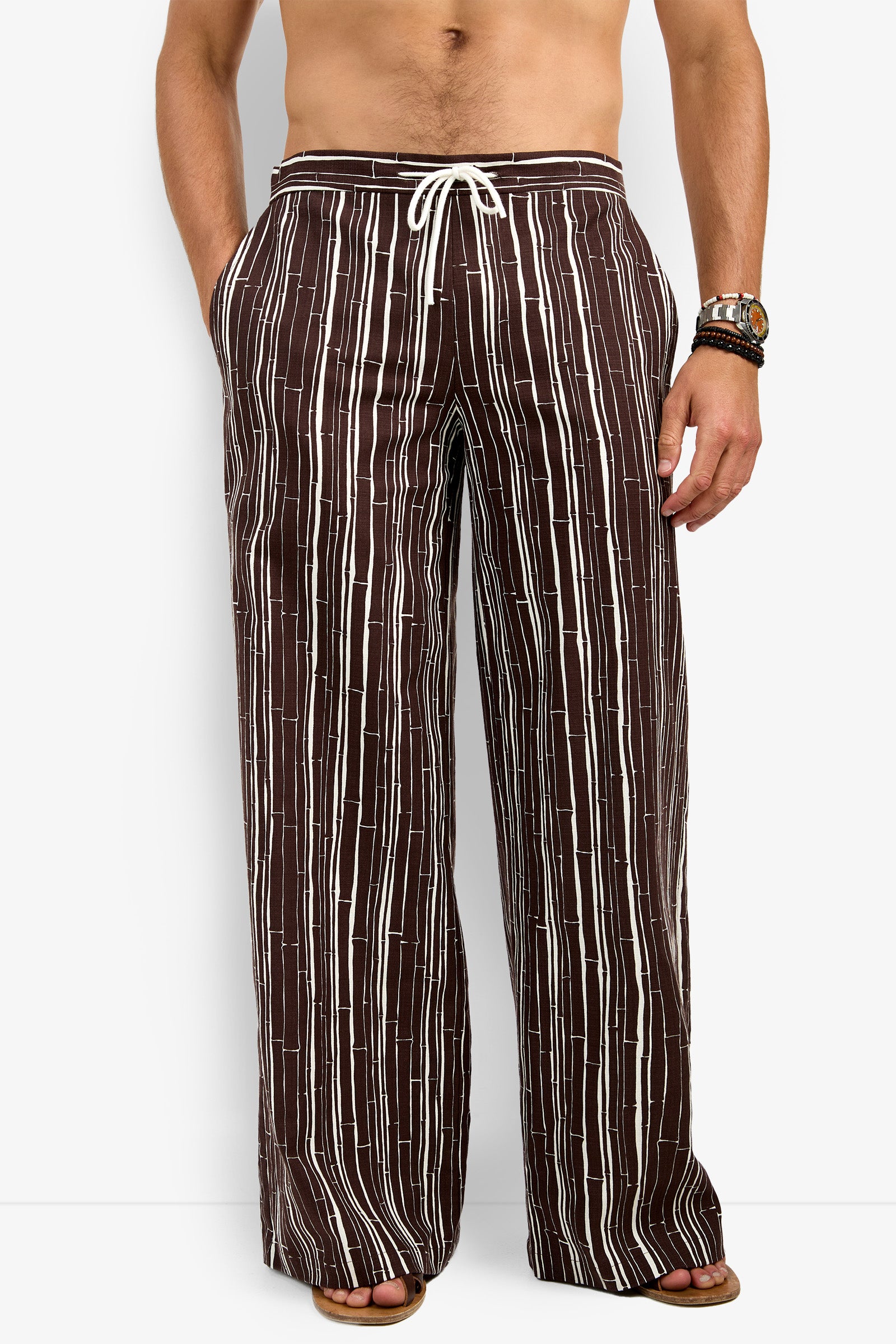 A man models the Wakiki Trouser—loose brown pants with vertical white stripes and a white drawstring waist—paired with sandals, shirtless, one hand in his pocket, bracelets and a watch on the other arm.