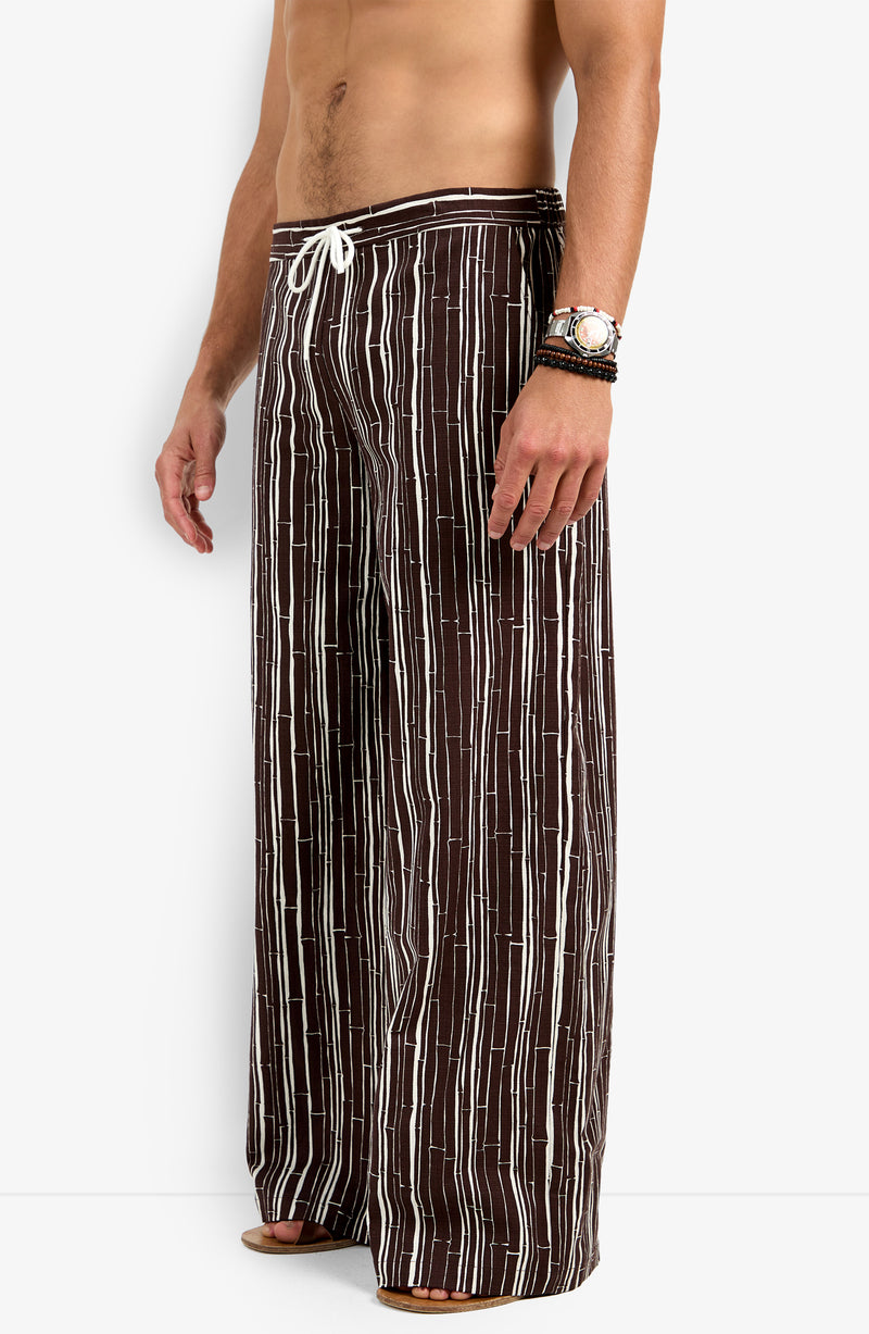 Model shown from mid-torso down wearing the Wakiki Trouser—loose, wide-leg brown pants with vertical white stripes and a white drawstring waist. The model is barefoot and accessorized with bracelets and a watch.