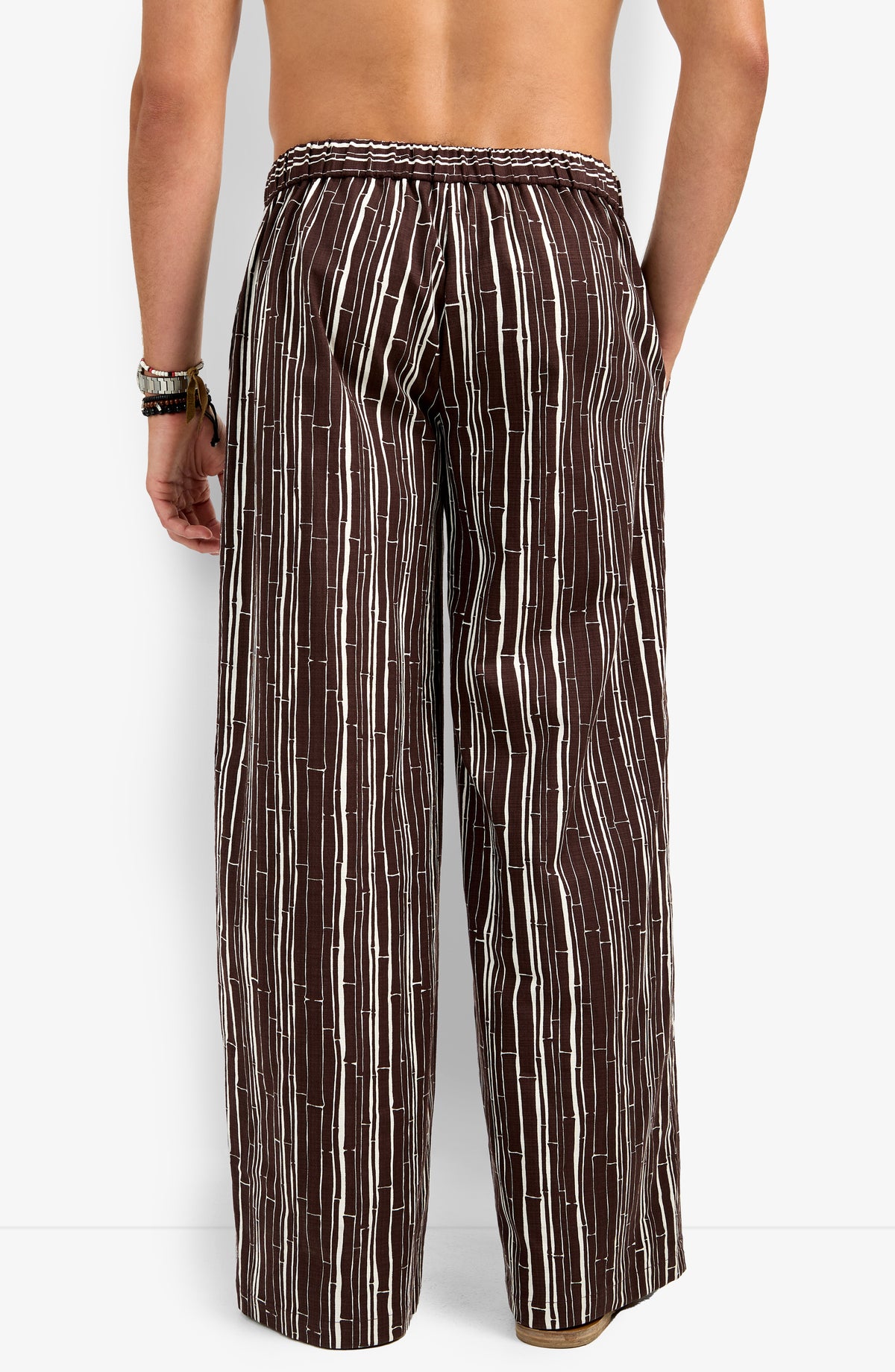 A person stands with their back to the camera, wearing the Wakiki Trouser—loose-fitting brown pants with vertical white stripes, an elastic waistband, and a relaxed fit. They are shirtless and wearing bracelets on one wrist.
