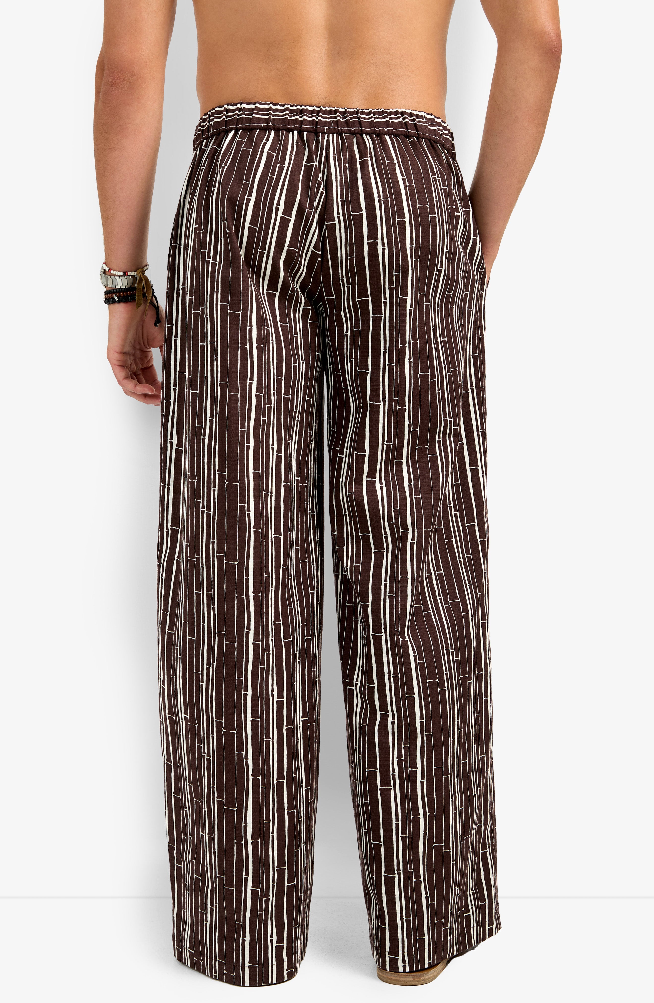 A person stands with their back to the camera, wearing the Wakiki Trouser—loose-fitting brown pants with vertical white stripes, an elastic waistband, and a relaxed fit. They are shirtless and wearing bracelets on one wrist.
