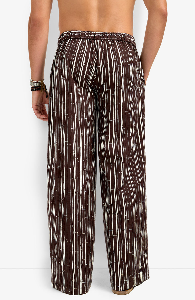 A person stands with their back to the camera, wearing the Wakiki Trouser—loose-fitting brown pants with vertical white stripes, an elastic waistband, and a relaxed fit. They are shirtless and wearing bracelets on one wrist.