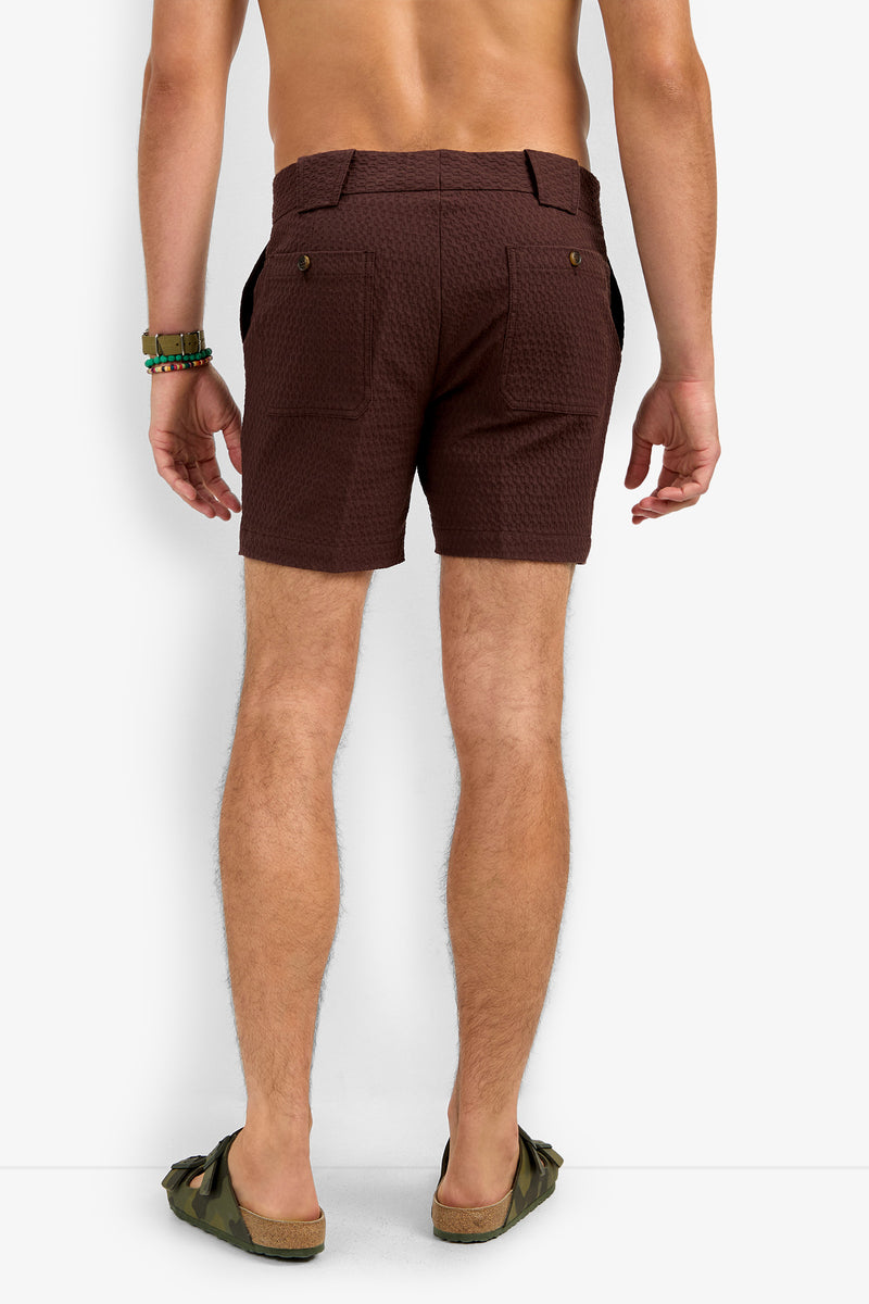 A man faces away from the camera, wearing the Randy Short—textured brown shorts—bracelets on his left wrist, and green sandals. His upper body is bare, with a plain white background.