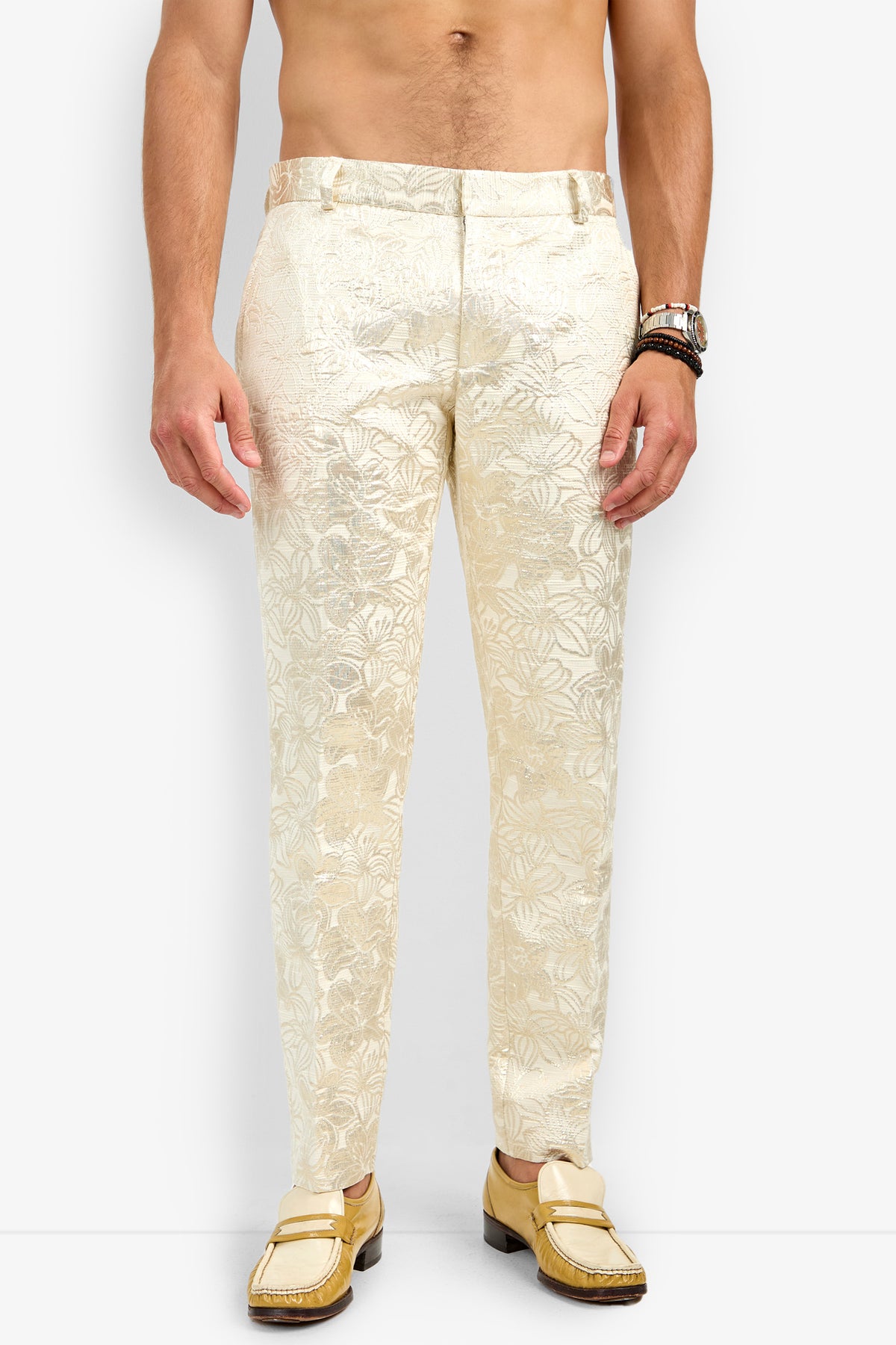 A shirtless man models the Clyde Slim Trouser—cream-colored, floral-patterned dress pants—with gold loafers against a white background. Only his body from shoulders to feet is visible, accessorized with a watch and bracelets on his left wrist.