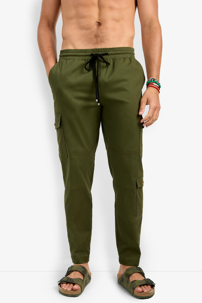 A man wears the San Pedro Jogger in olive green with a drawstring waist and side pockets, paired with olive sandals. He is shirtless, has bracelets on his right wrist, and stands against a plain white background.