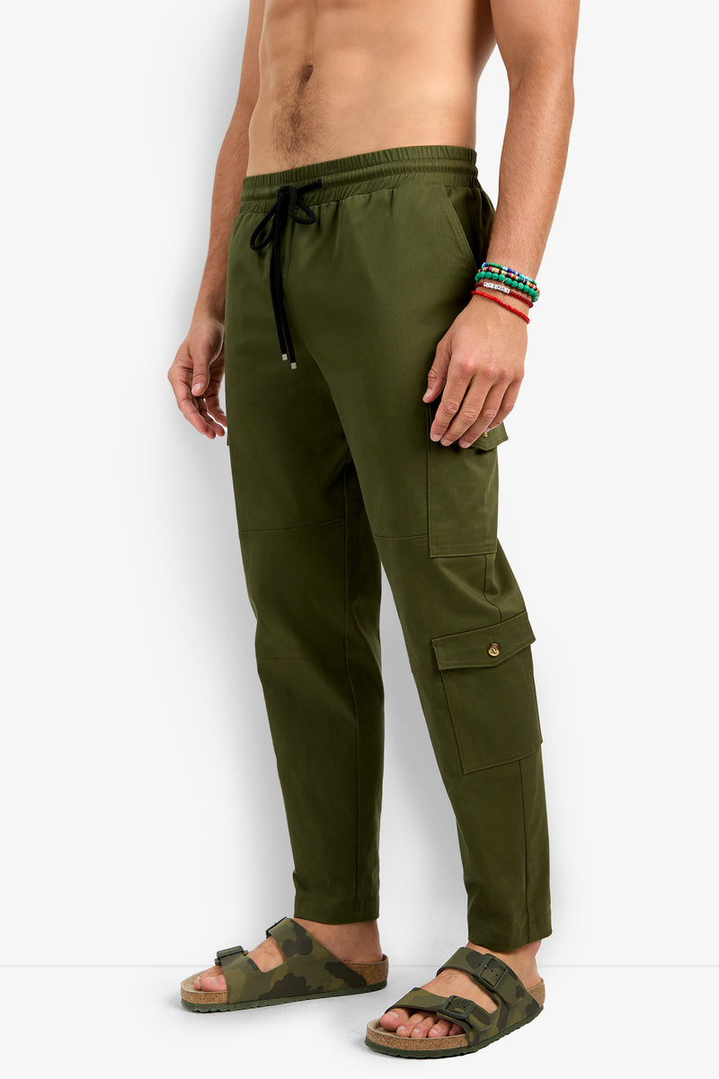 A shirtless man models the San Pedro Jogger in olive green, featuring a drawstring waist. He accessorizes with beaded bracelets and camouflage sandals while standing against a plain white background.