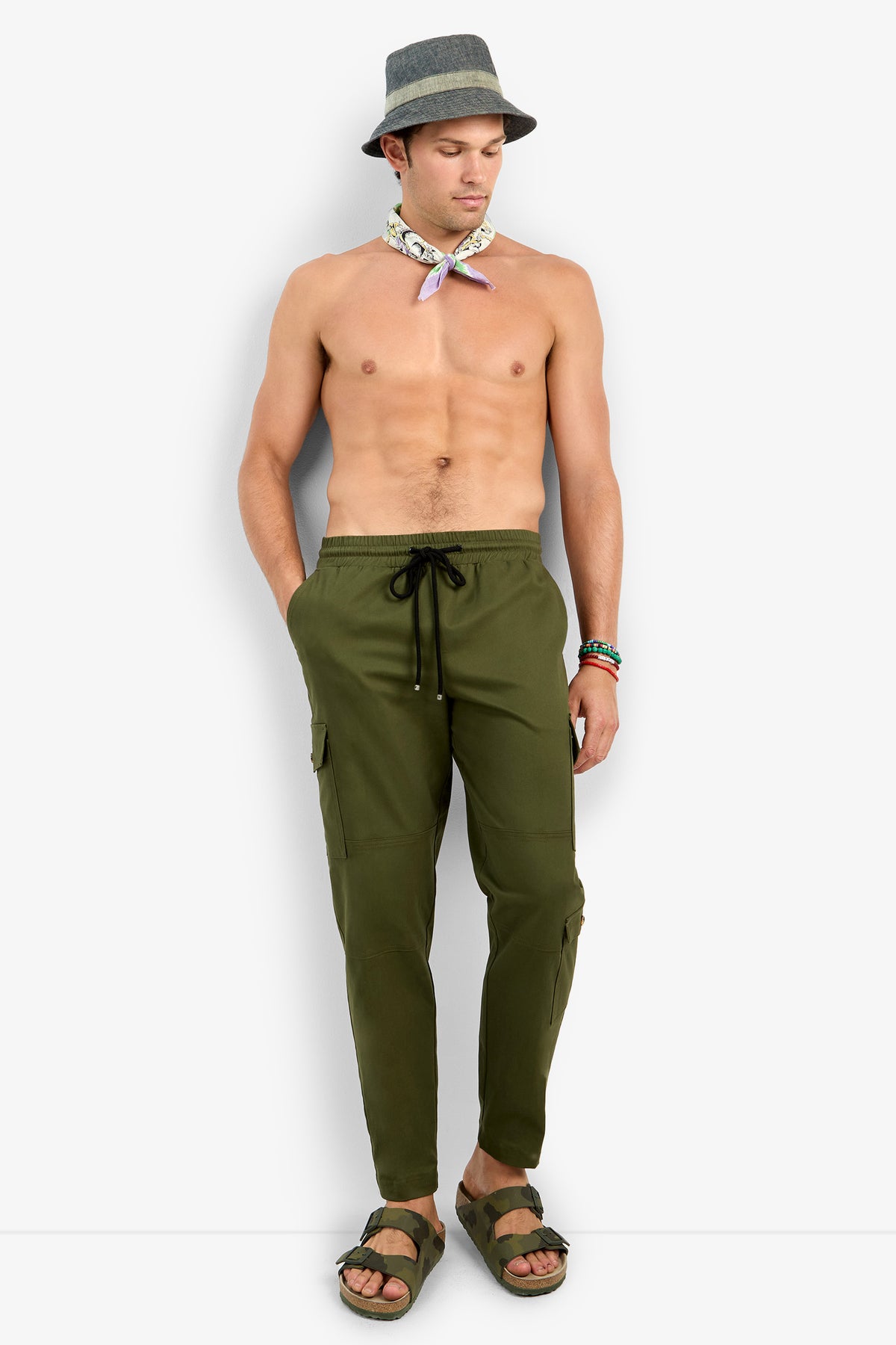 A shirtless man wearing olive green San Pedro Jogger pants, a gray hat, a light neck scarf, and green sandals stands against a plain white background with his hands in his pockets, looking down.