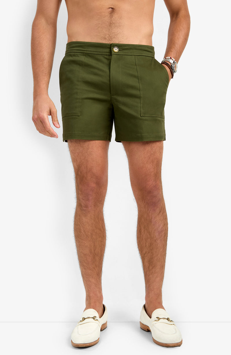 A man wears the Will Rogers Short in olive green and cream loafers against a white background. His torso is bare, a watch adorns his left wrist, and his hands rest casually by his sides.