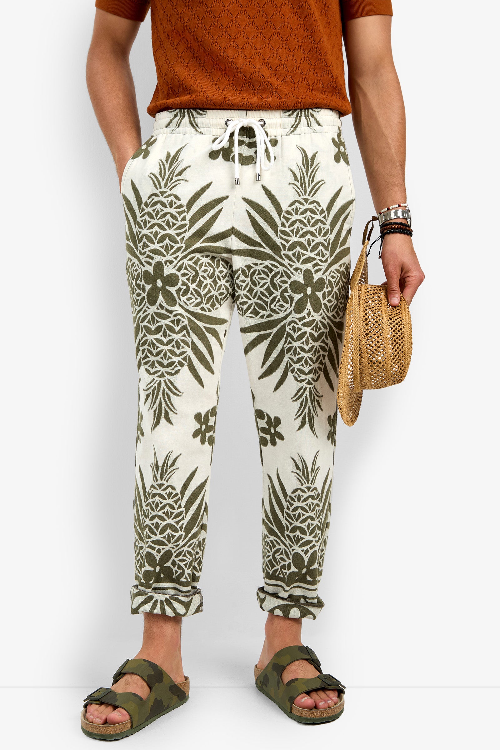 A person wears the Felipe Drawstring Pant—white joggers with green pineapple and leaf print—paired with a rust shirt, green camo sandals, bracelets, and a small woven straw bag. Hands are tucked in the pant pockets.