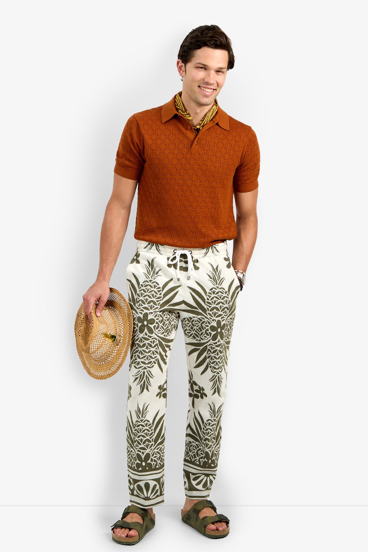 A man stands smiling, wearing orange short-sleeve knit polo, Felipe Drawstring Pant in white and green pattern, green sandals, a yellow patterned neck scarf, and holding a straw hat in one hand.