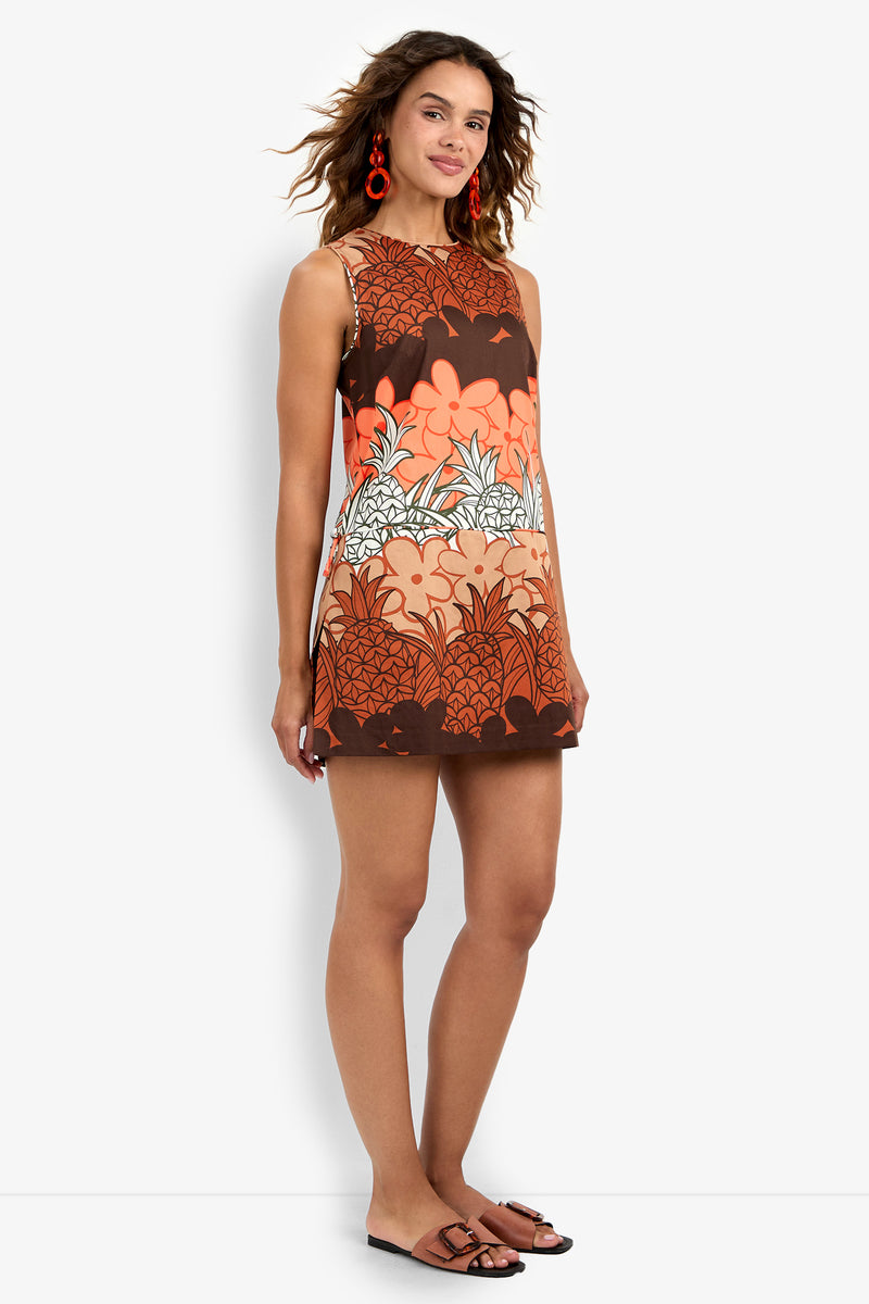 A woman stands and smiles, wearing the sleeveless Deniz Romper with a bold orange, brown, and white floral pattern, paired with brown sandals and red circular earrings.