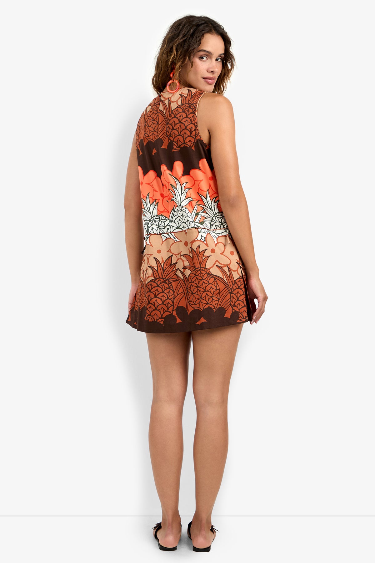 A woman faces away, turning to smile at the camera. She wears the sleeveless Deniz Romper with a bold orange, brown, and cream floral print and black sandals against a plain white background.