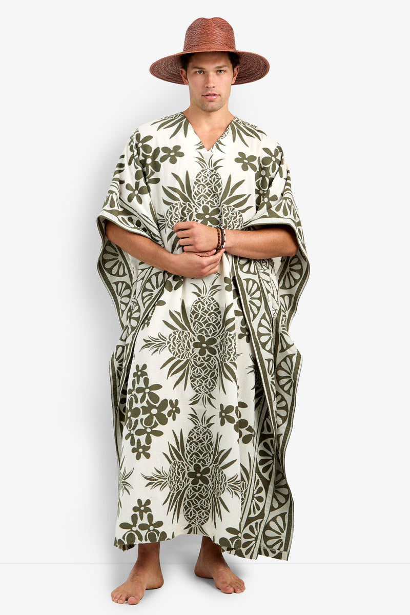 A barefoot person stands facing forward, wearing the Wailea Caftan—a loose cream robe with green tropical florals—and a wide-brimmed brown hat, hands clasped in front.