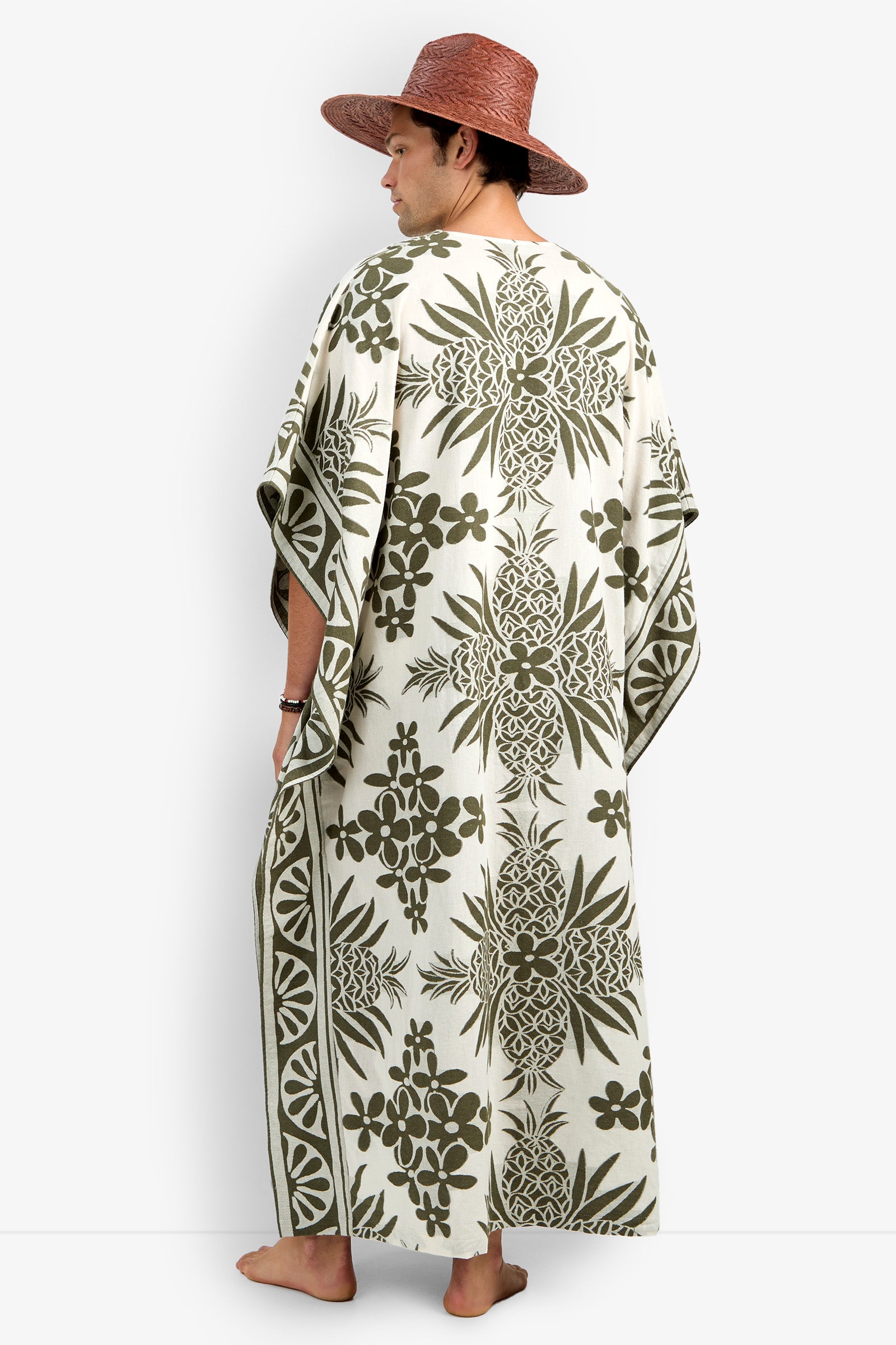 A person wearing the Wailea Caftan, a cream-colored robe with green tropical pineapple and floral prints, paired with a wide-brimmed brown hat, stands barefoot and faces away from the camera.