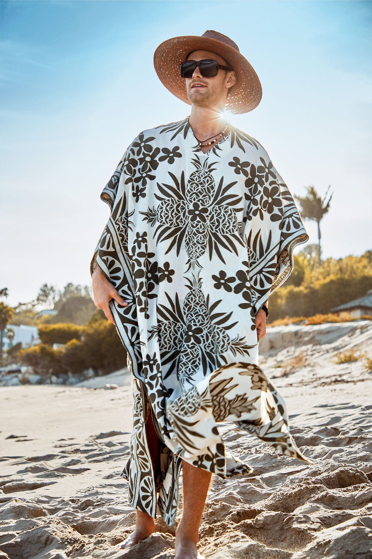 A person wearing the Wailea Caftan stands barefoot on a sandy beach, surrounded by greenery and a clear blue sky.