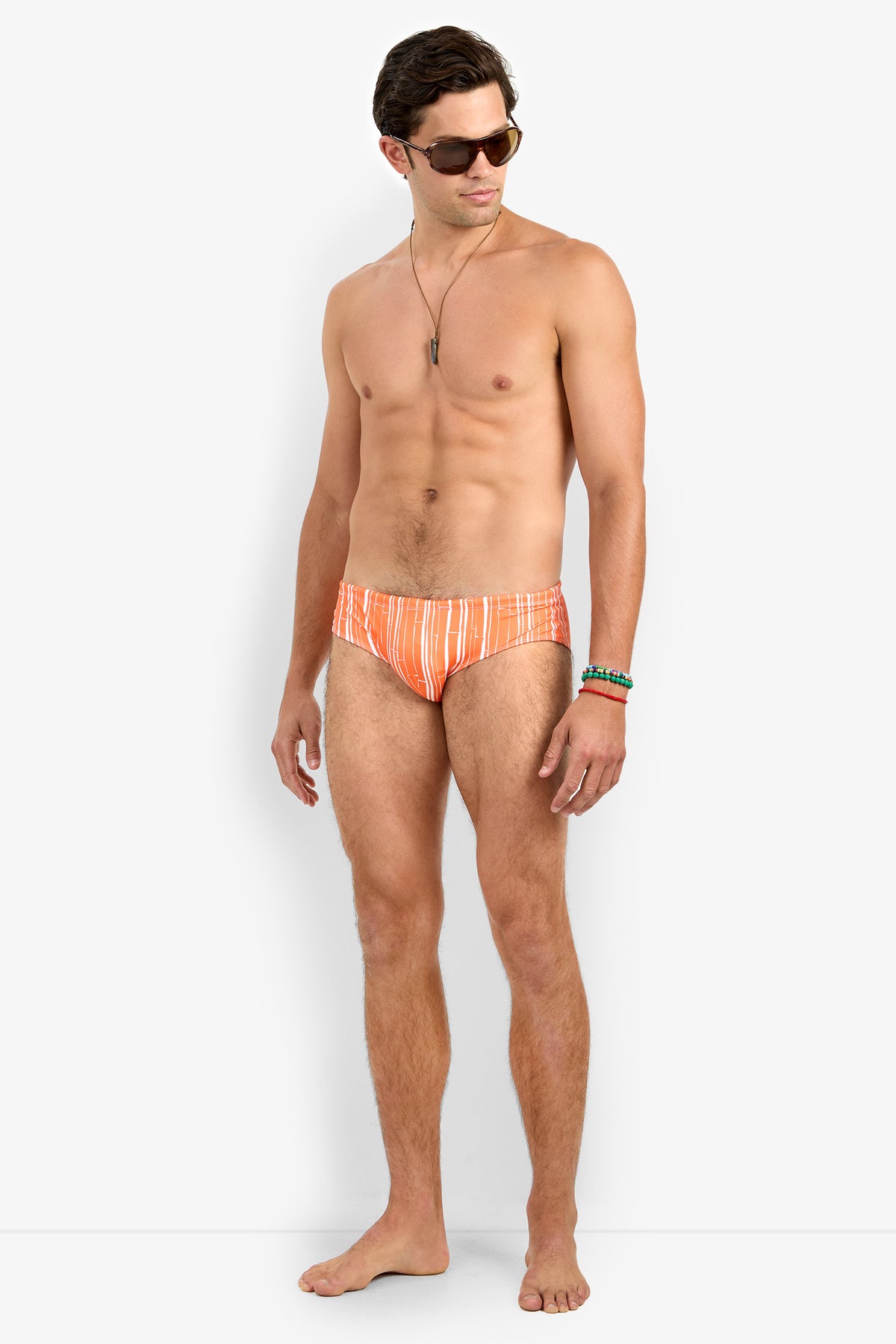 A man wearing sunglasses, a necklace, and bracelets models the Bondi Swim Brief—an orange striped swim brief—standing barefoot against a plain white background and looking to his left.