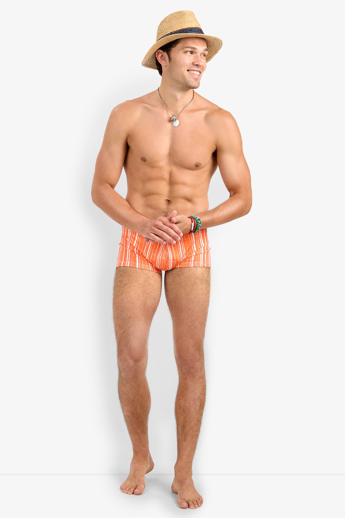 A smiling man stands barefoot against a plain background, looking to the side with his hands clasped, wearing a straw hat, a necklace, bracelets, and Sorrento Swim Trunk in orange and white stripes.