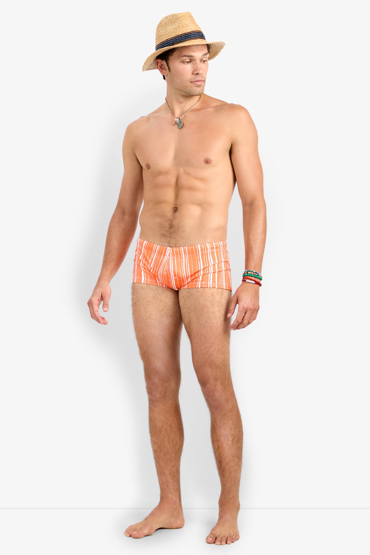 A man stands barefoot against a plain white background, wearing a straw hat, colorful bracelets, a necklace, and the Sorrento Swim Trunk—an orange and white patterned swimsuit.