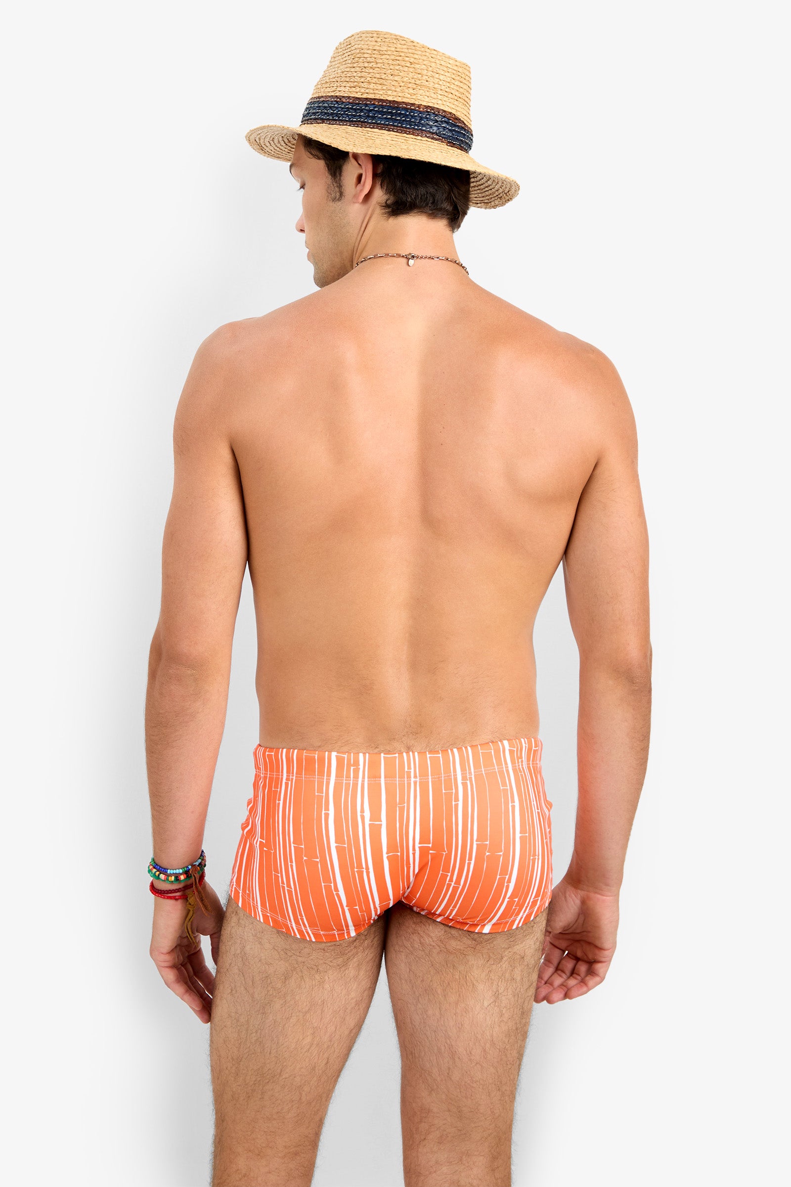 A man in Sorrento Swim Trunk, an orange and white striped swimsuit, stands shirtless with his back to the camera, wearing a straw hat and colorful bracelets. The background is plain white.