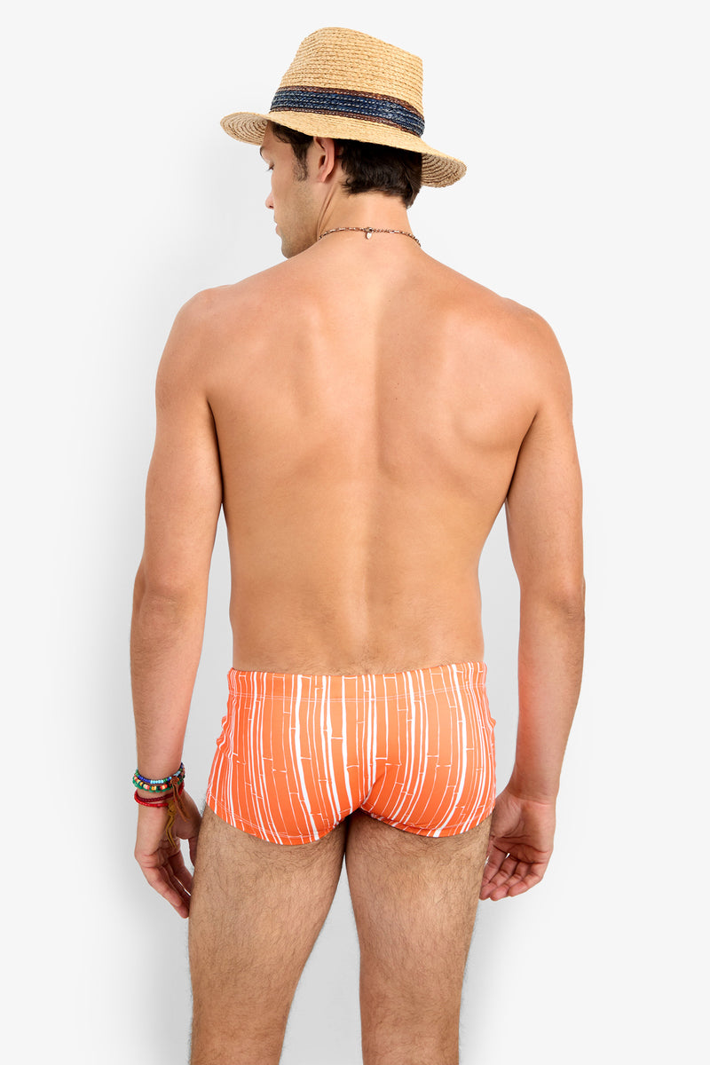 A man in Sorrento Swim Trunk, an orange and white striped swimsuit, stands shirtless with his back to the camera, wearing a straw hat and colorful bracelets. The background is plain white.
