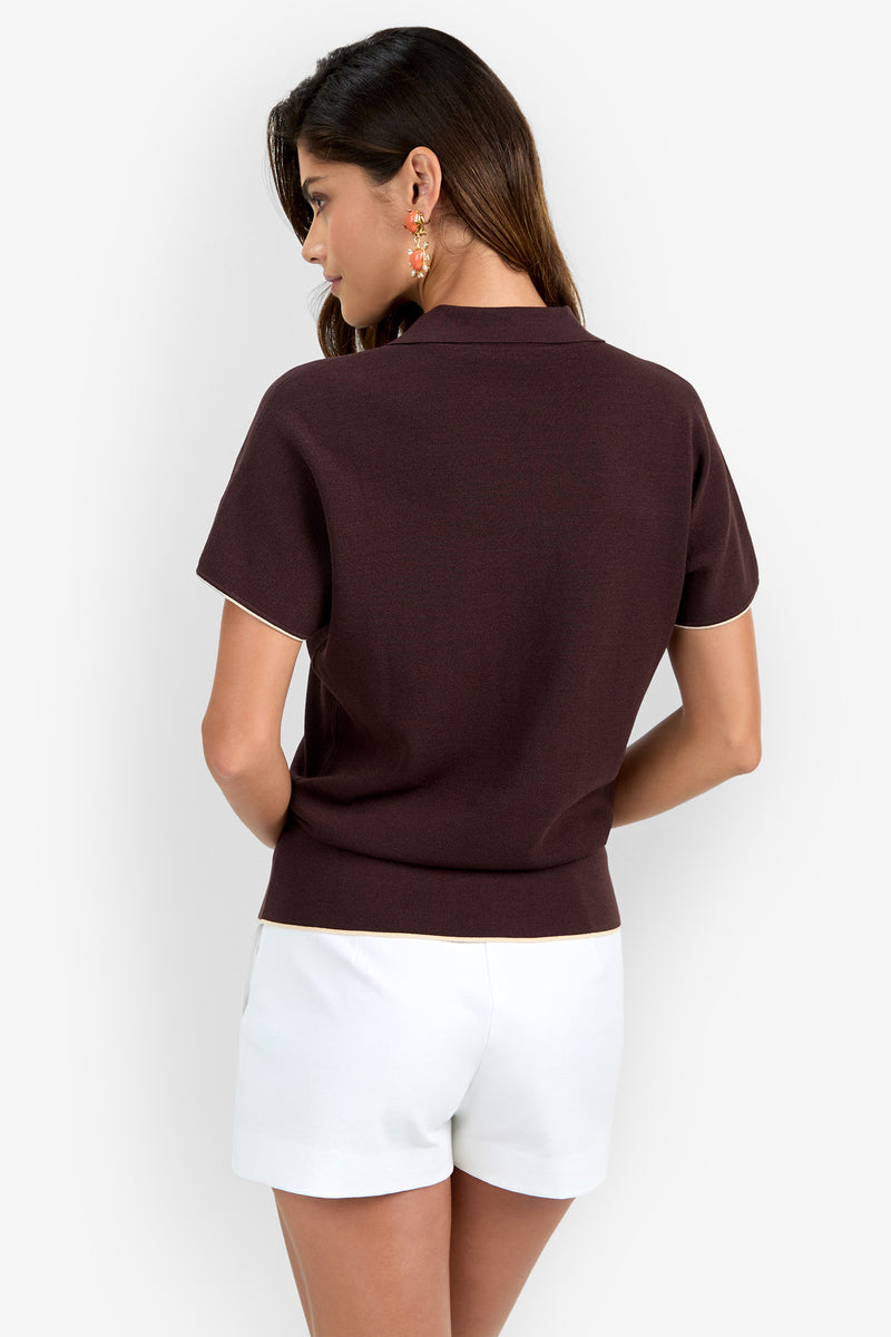 A woman with long brown hair and gold earrings stands against a plain light background, wearing the Hilo Pullover—a short-sleeve dark brown top—paired with white shorts.