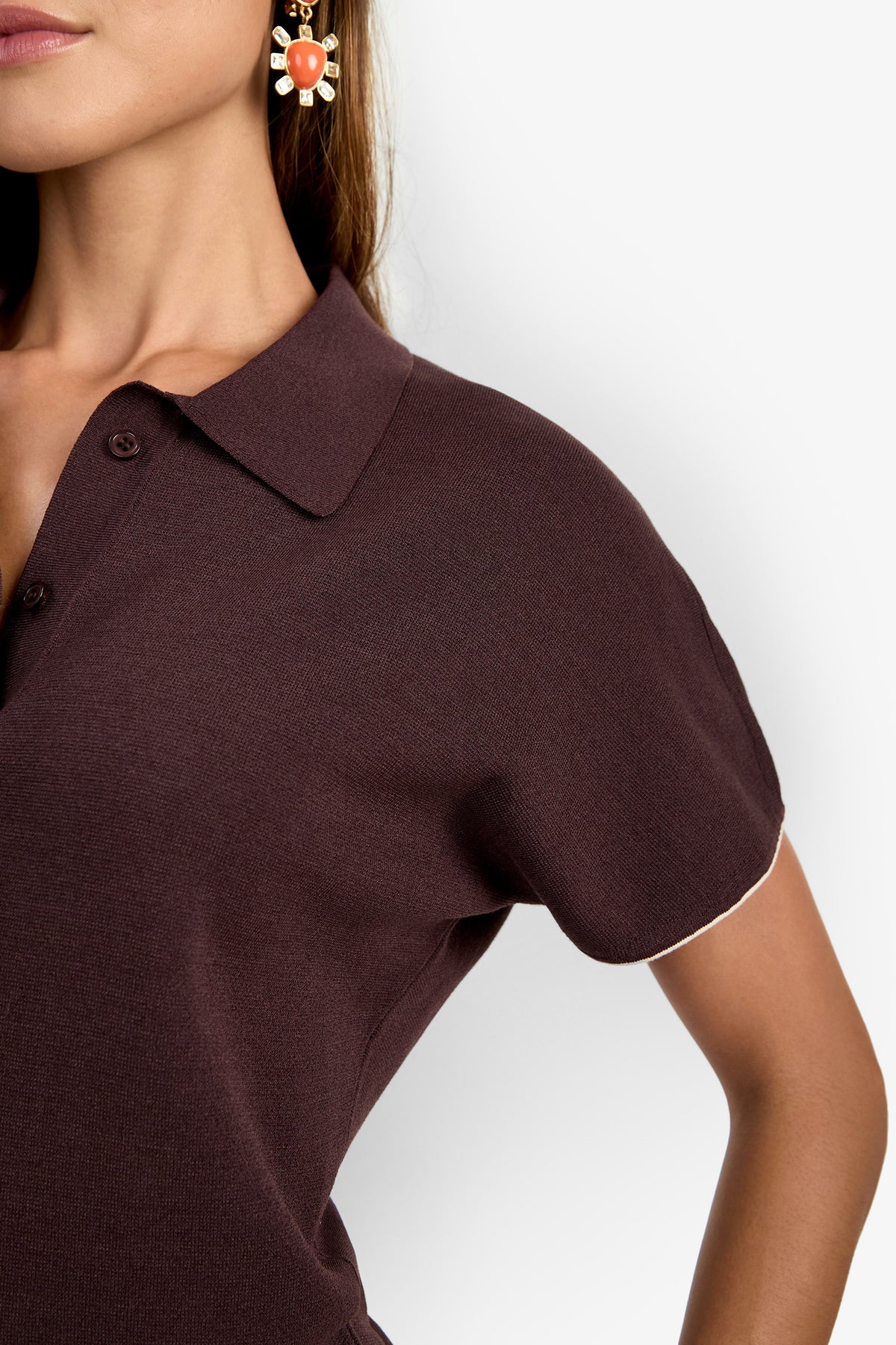 A woman wears the Hilo Pullover, a maroon collared shirt with short sleeves, paired with a decorative earring as she poses with one hand on her hip against a plain white background.
