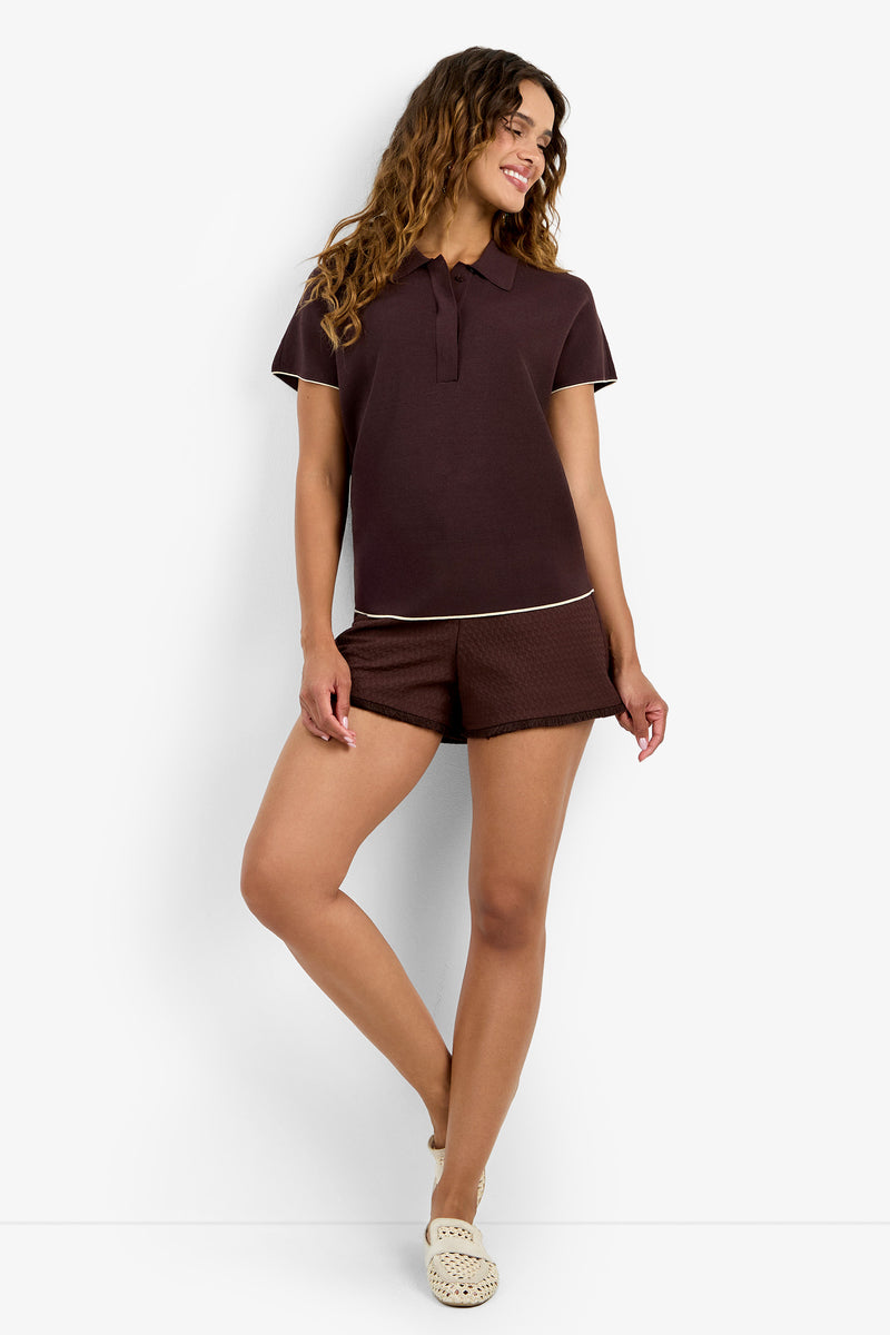 A woman smiles and stands with one leg crossed, wearing the HILO PULLOVER in CAROB/BONITA BEIGE against a white background.
