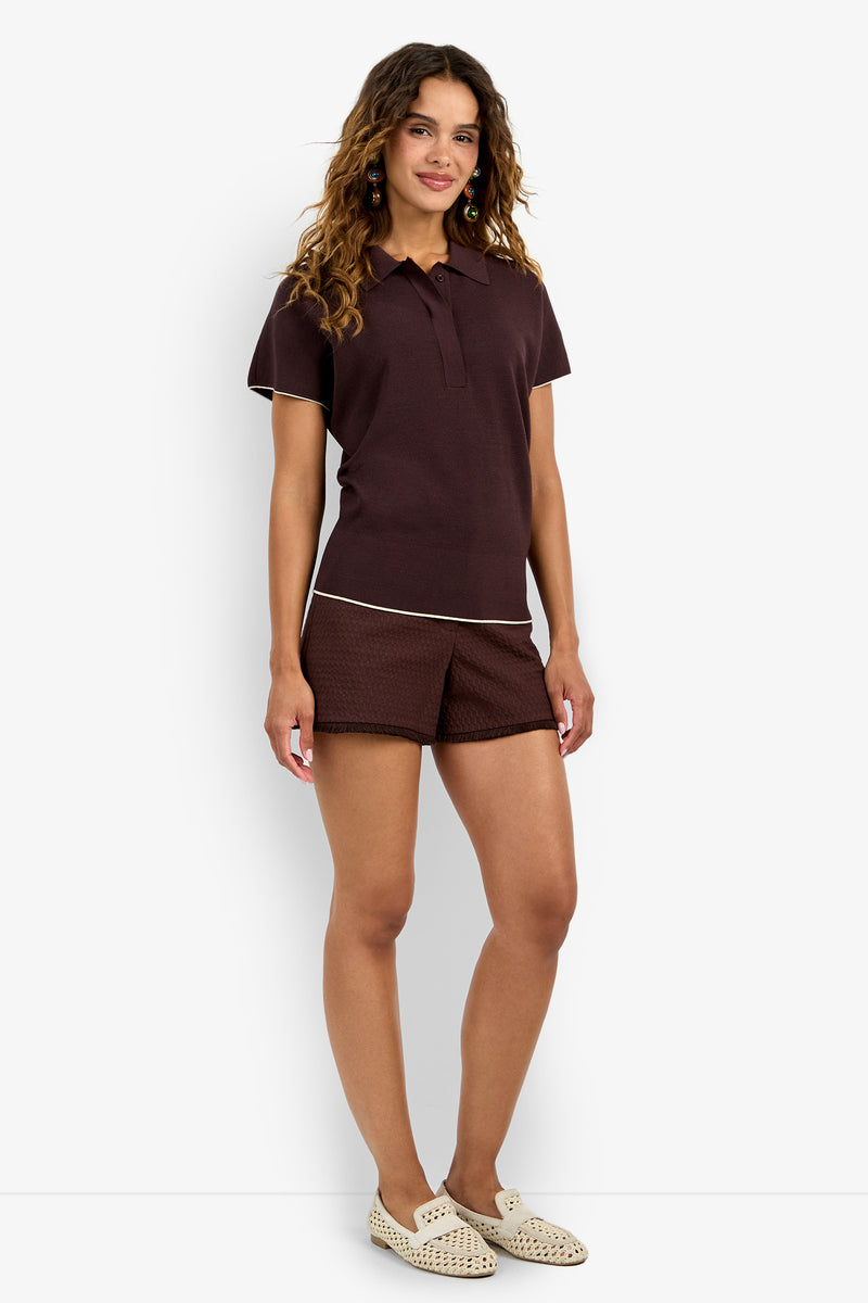 A woman with wavy brown hair and a relaxed expression stands against a white background, wearing the Hilo Pullover in dark brown, matching patterned shorts, and cream-colored slip-on shoes.