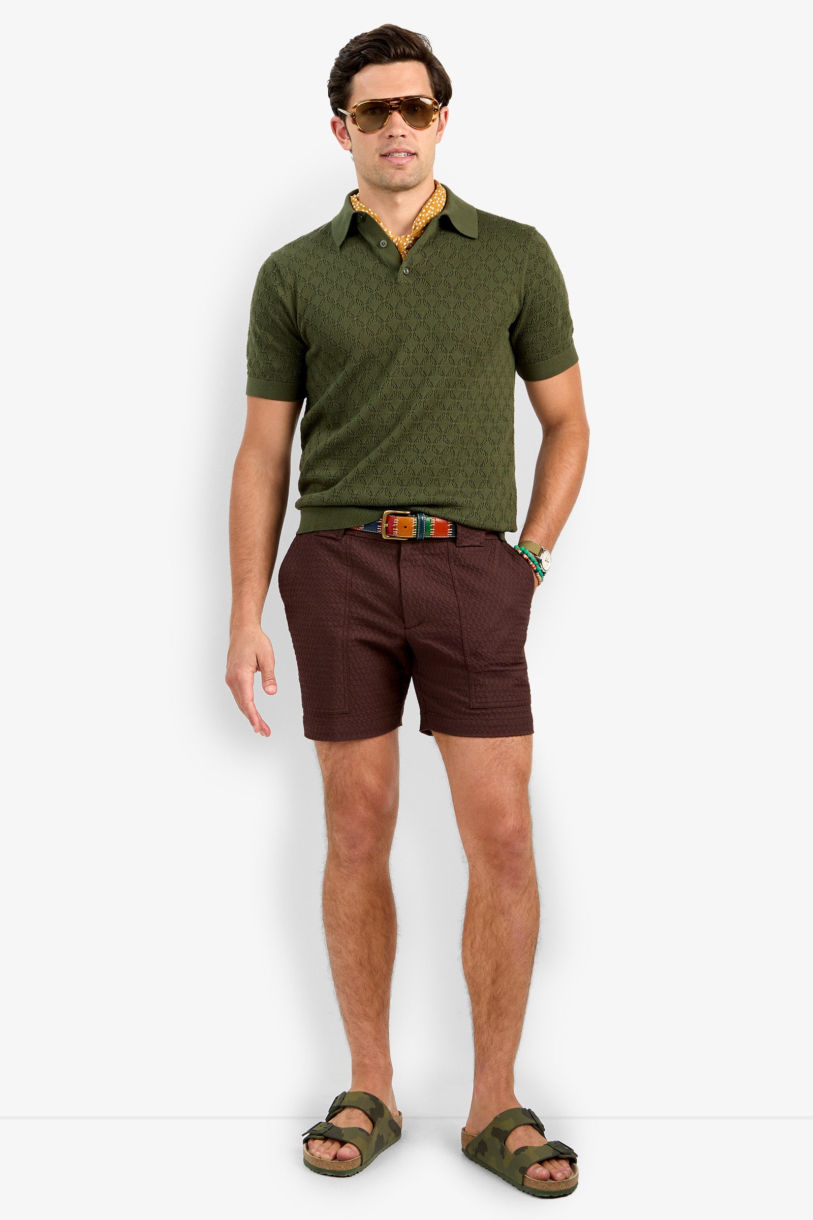 A man wearing sunglasses, the Waylen Short Sleeve Polo in olive green, brown shorts, a colorful belt, and green sandals stands facing forward against a plain white background.