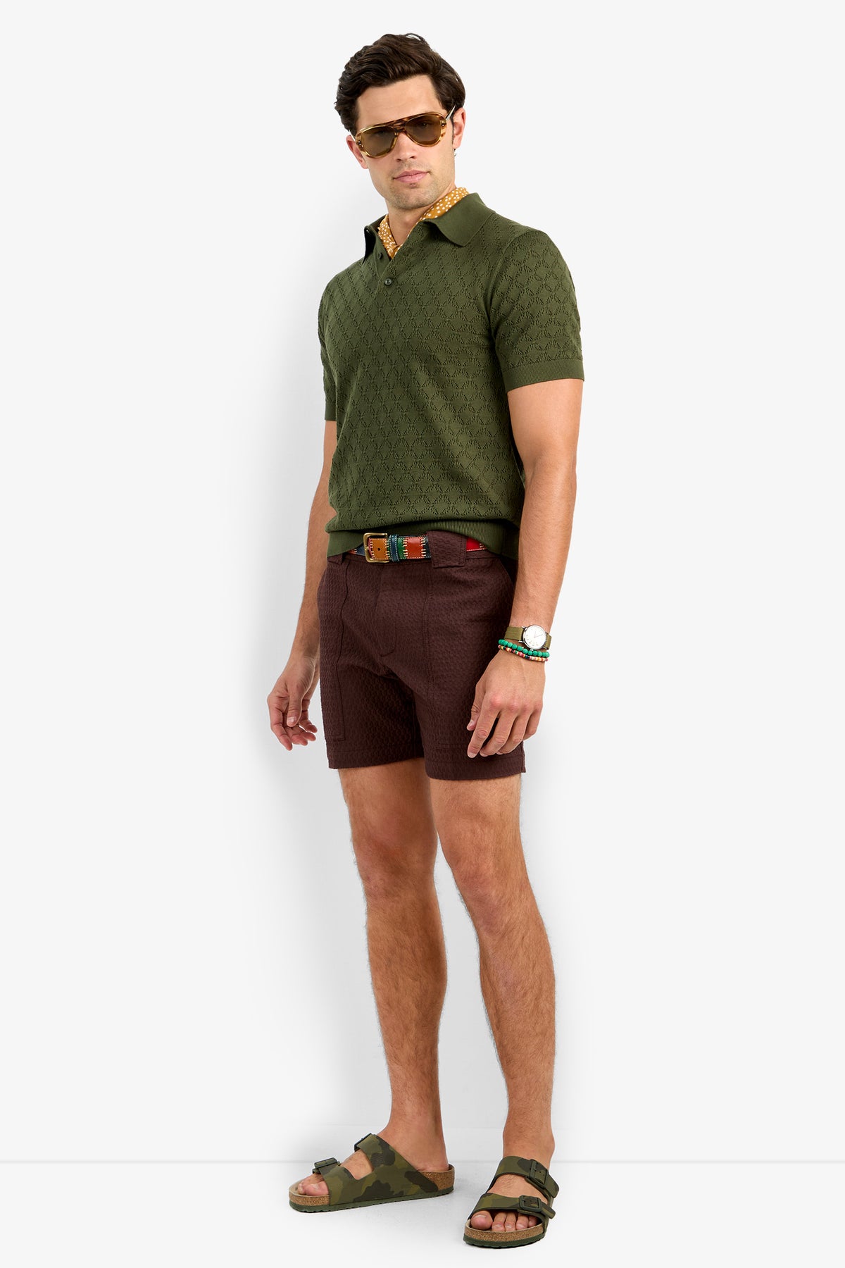 A man in sunglasses, the Waylen Short Sleeve Polo in olive green, patterned brown shorts, a colorful belt, watch, and green camo sandals stands against a plain white background.