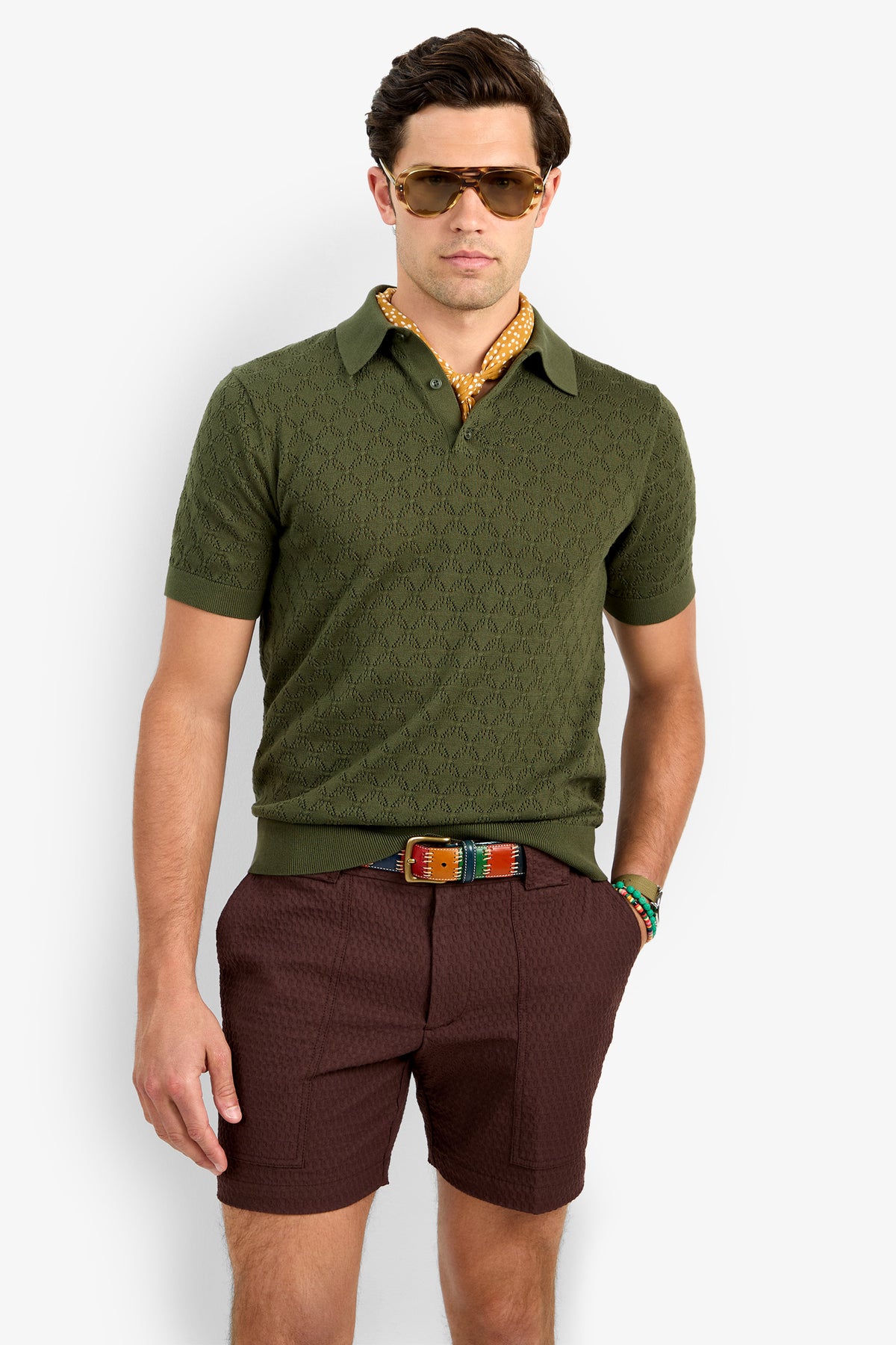 A man in sunglasses, the green Waylen Short Sleeve Polo, patterned neck scarf, brown shorts, and a colorful belt stands against a plain light background with one hand in his pocket.